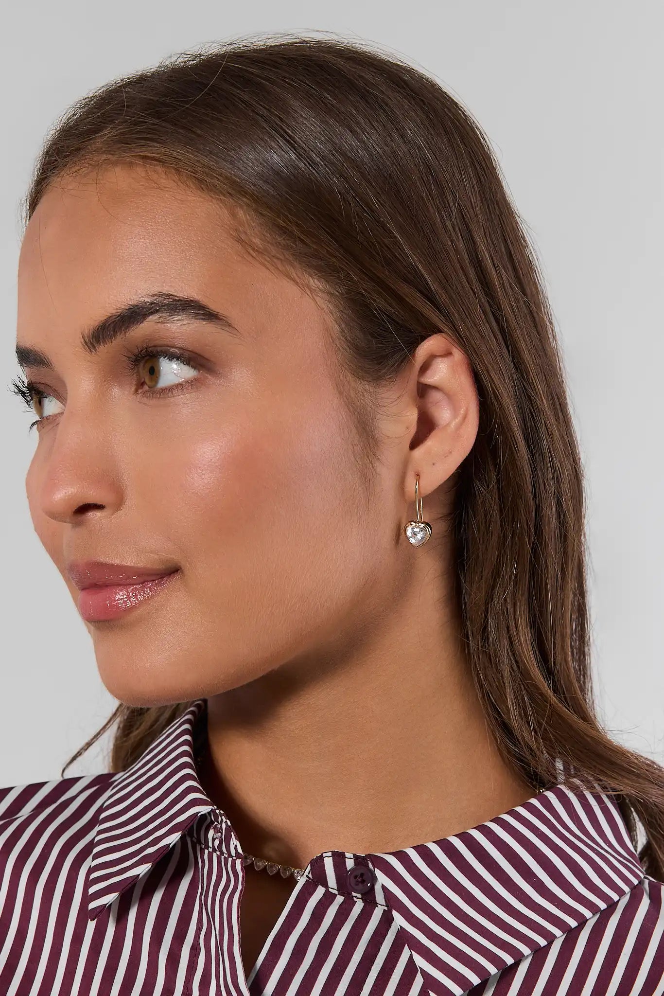 A close-up side profile of a woman with brown hair wearing a maroon and white striped shirt, showcasing a small gold hoop earring with a heart-shaped clear gemstone.