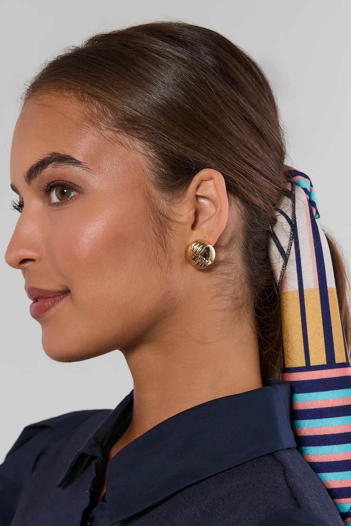 A close-up side profile of a woman wearing a textured gold dome earring, with her hair tied back and a striped scarf visible behind her neck, against a neutral gray background.