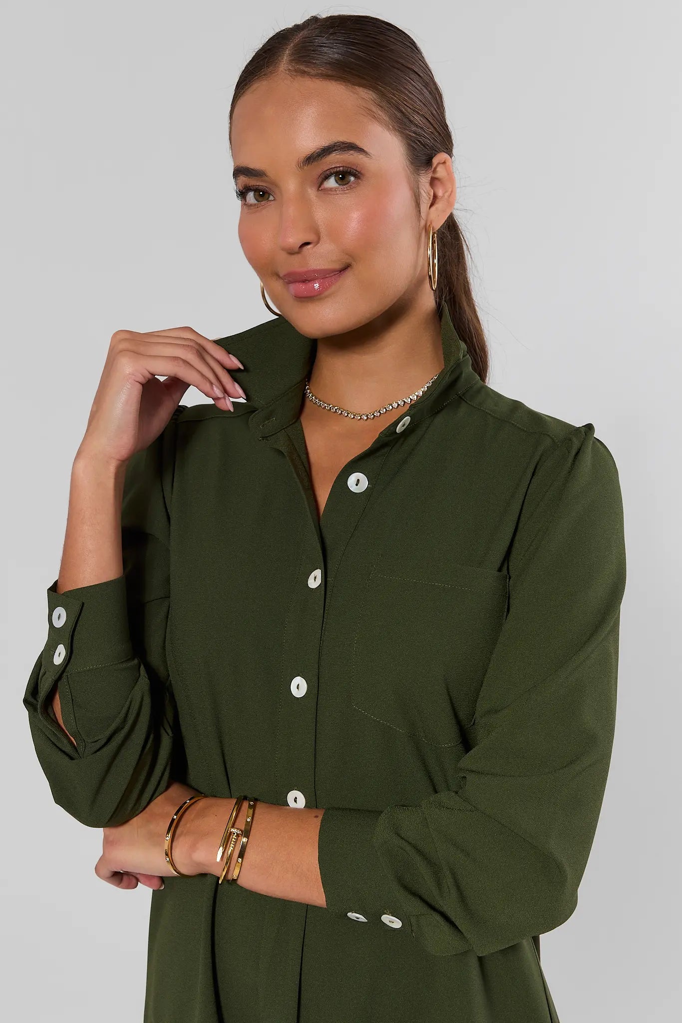 A woman wearing an olive green long-sleeve button-up shirt with white buttons, shown from the chest up against a light gray studio background.