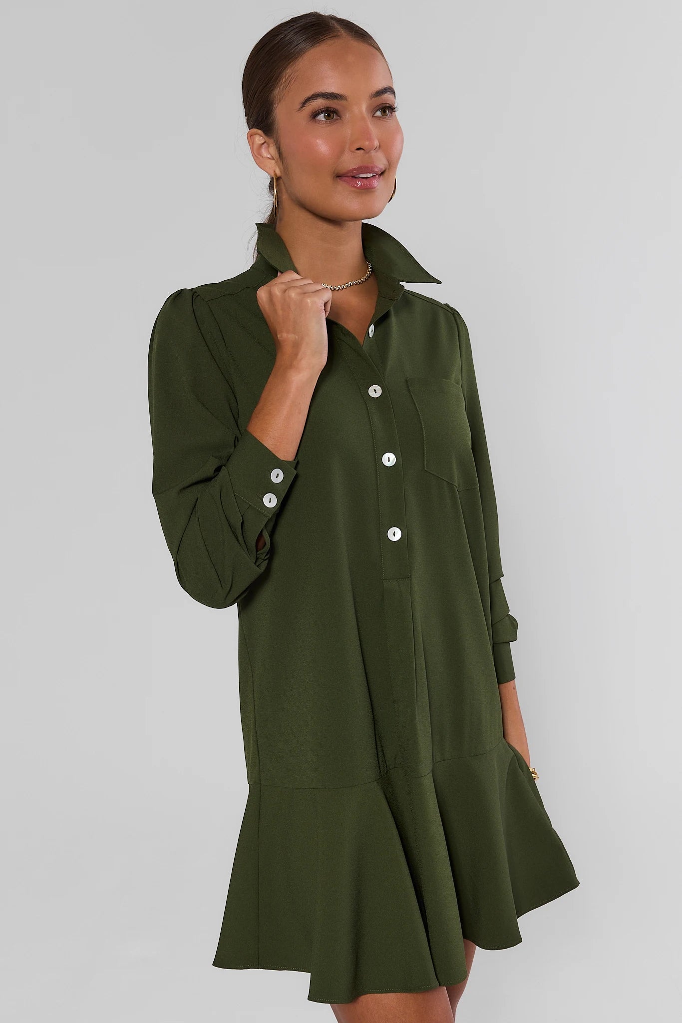 A woman modeling an olive green long-sleeve shirt dress with a button-down front and flared hem against a plain light gray studio background.