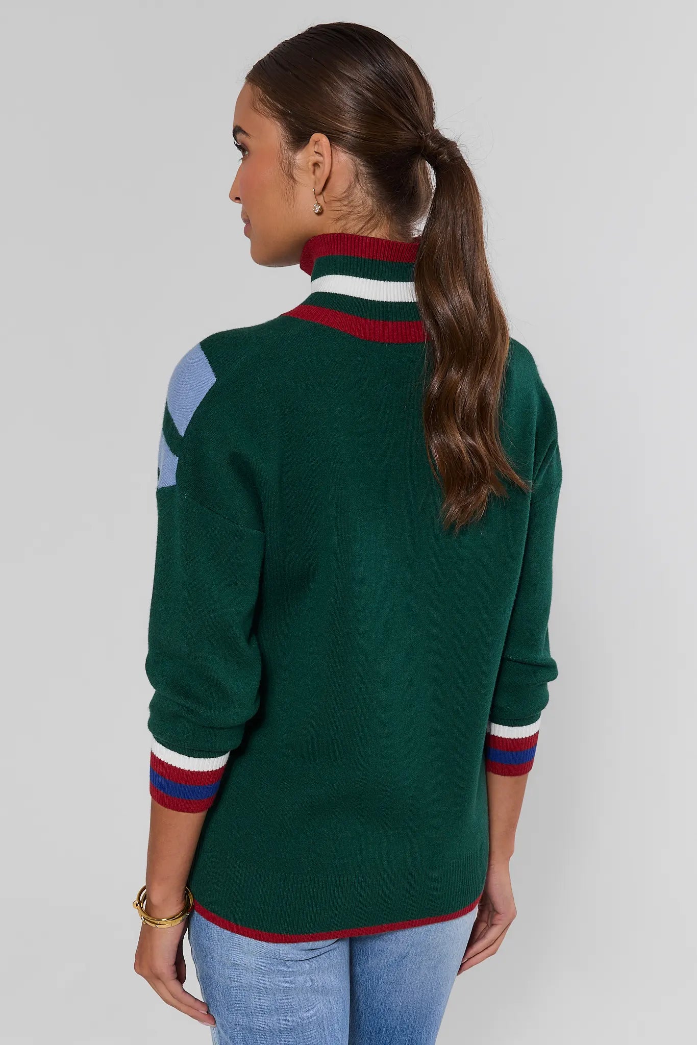 A woman shown from the back wearing a dark green knit sweater with red, white, and light blue color-blocked details on the collar and sleeves, paired with light blue jeans against a light gray studio background.
