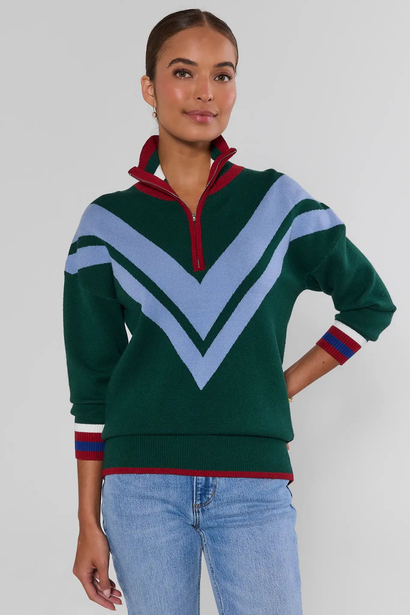 A woman modeling a green half-zip sweater with a light blue chevron pattern and red and blue striped cuffs, paired with light blue jeans, against a plain light gray background.