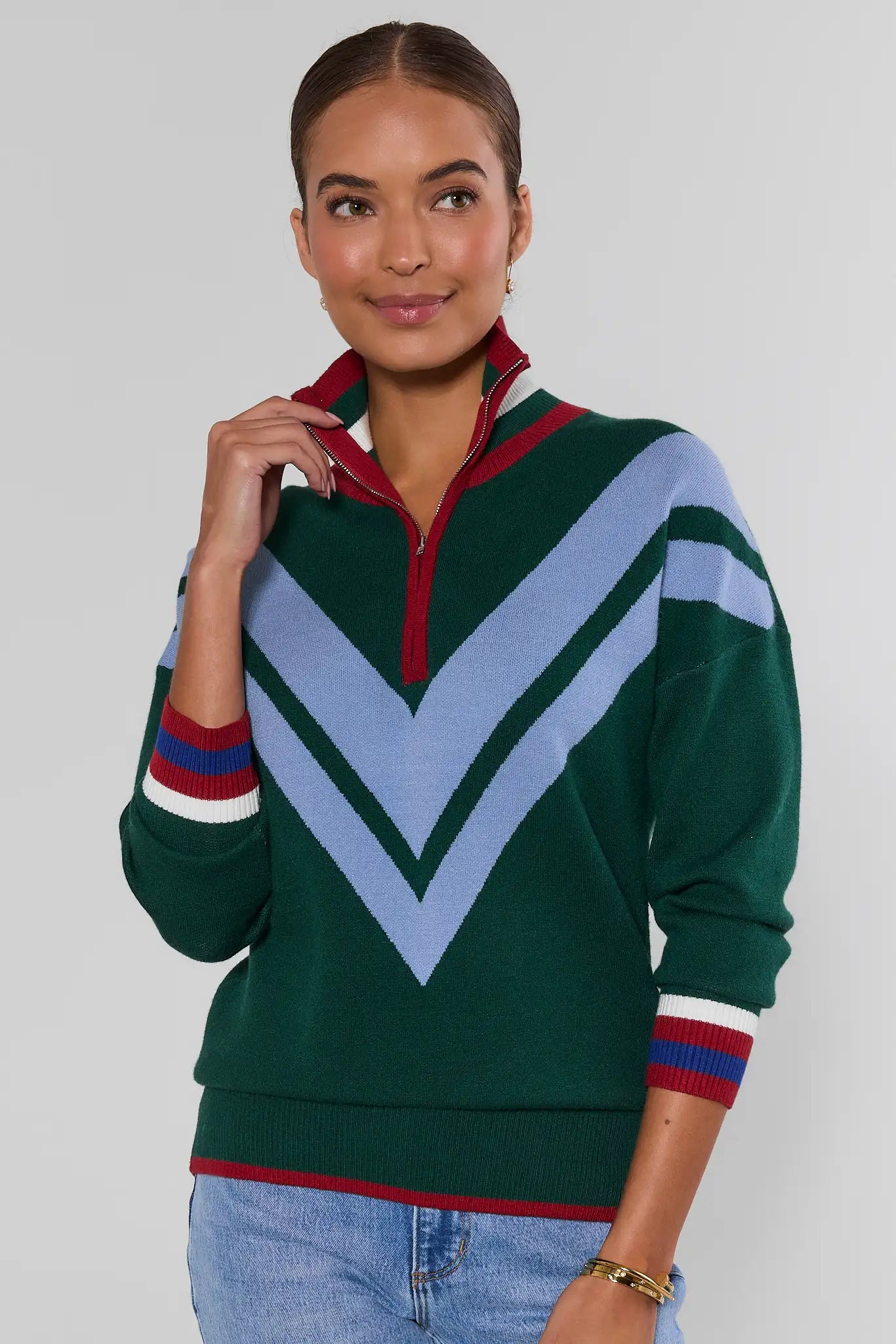 A woman wearing a green quarter-zip sweater with a large light blue chevron pattern on the front, red and white striped cuffs, and a red collar, paired with light blue jeans against a light gray studio background.