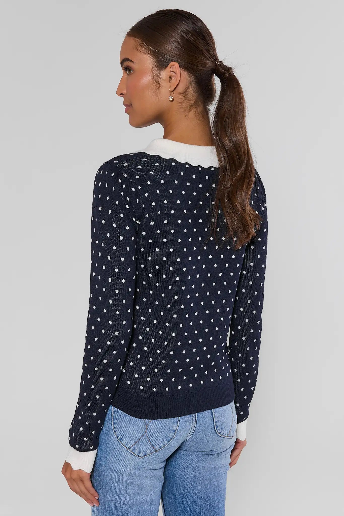 A woman shown from the back wearing a navy blue long-sleeve sweater with white polka dots and a white ruffled collar, paired with light blue jeans, against a light gray studio background.