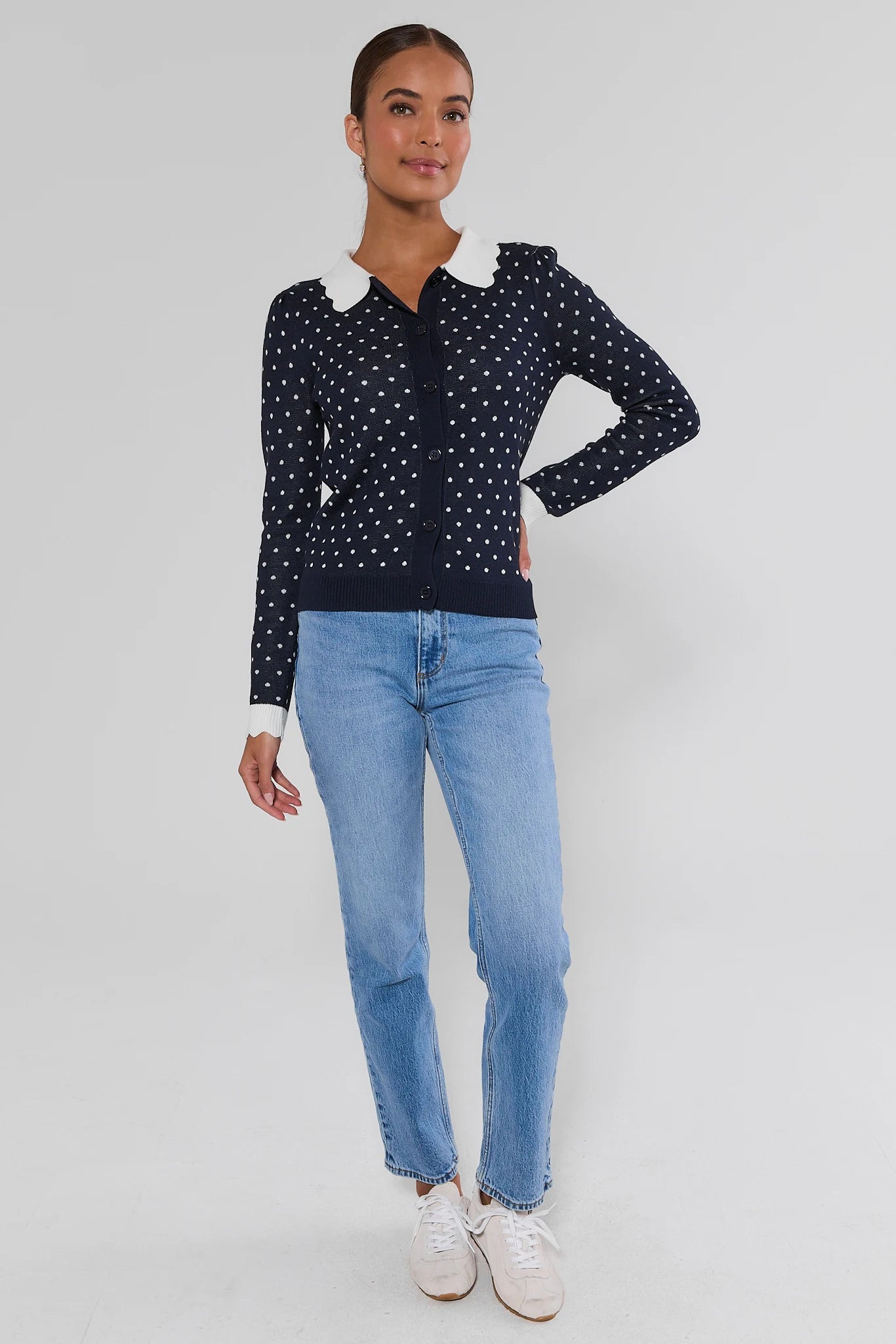 A woman modeling a navy blue long-sleeve knit polo cardigan with white polka dots and a contrasting white collar, paired with light blue jeans and white sneakers, against a light gray studio background.