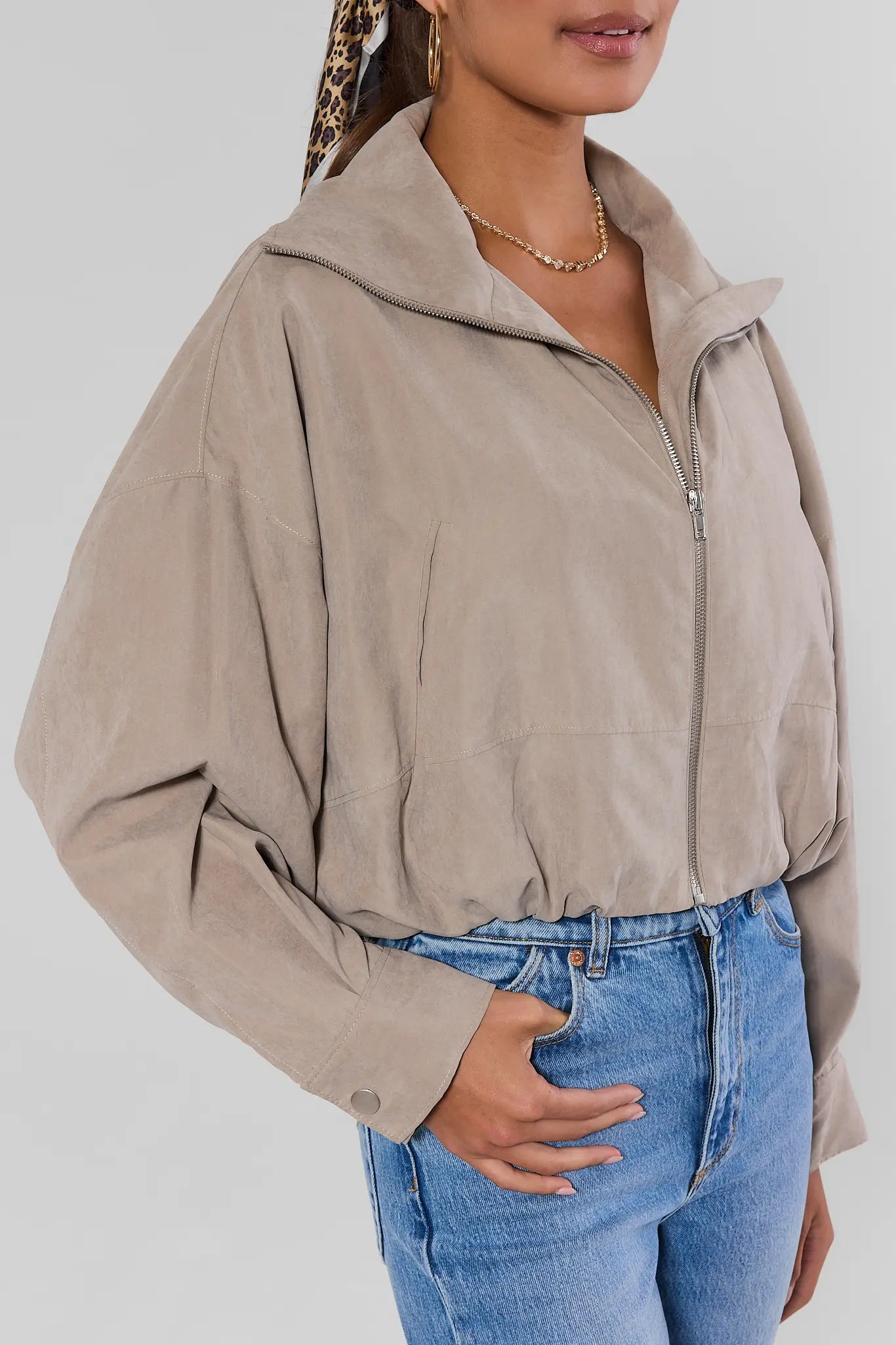 A woman wearing a taupe-beige cropped zip-up jacket with a high collar and wide sleeves, paired with blue jeans, against a light gray studio background.