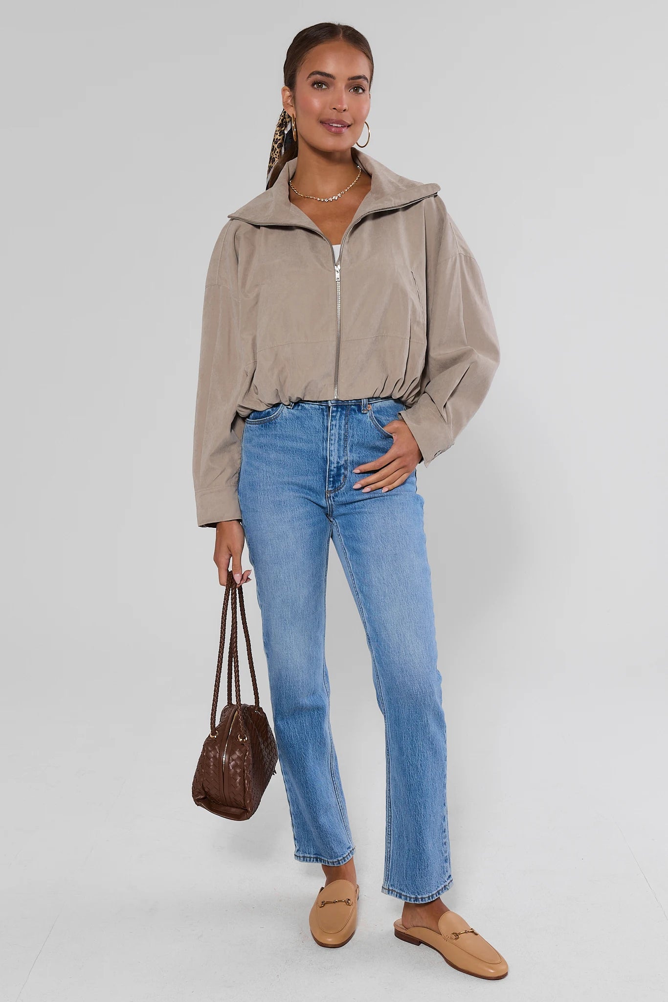 A woman modeling a beige cropped zip-up jacket with a high collar, paired with light-wash blue jeans, tan loafers, and a brown handbag, against a light gray studio background.