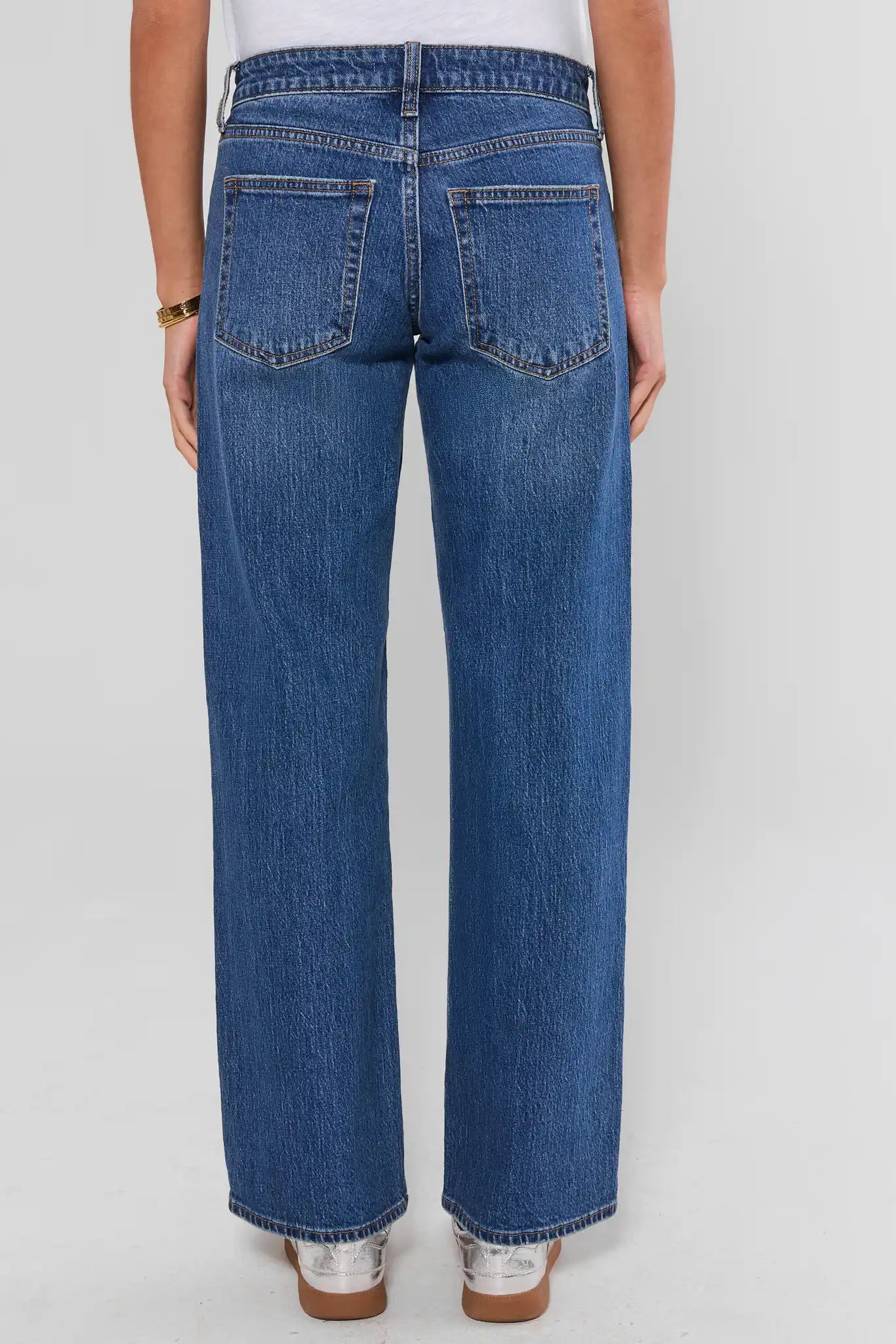 Rear view of a person wearing blue wide-leg jeans with a white top and metallic shoes, standing against a light gray studio background.
