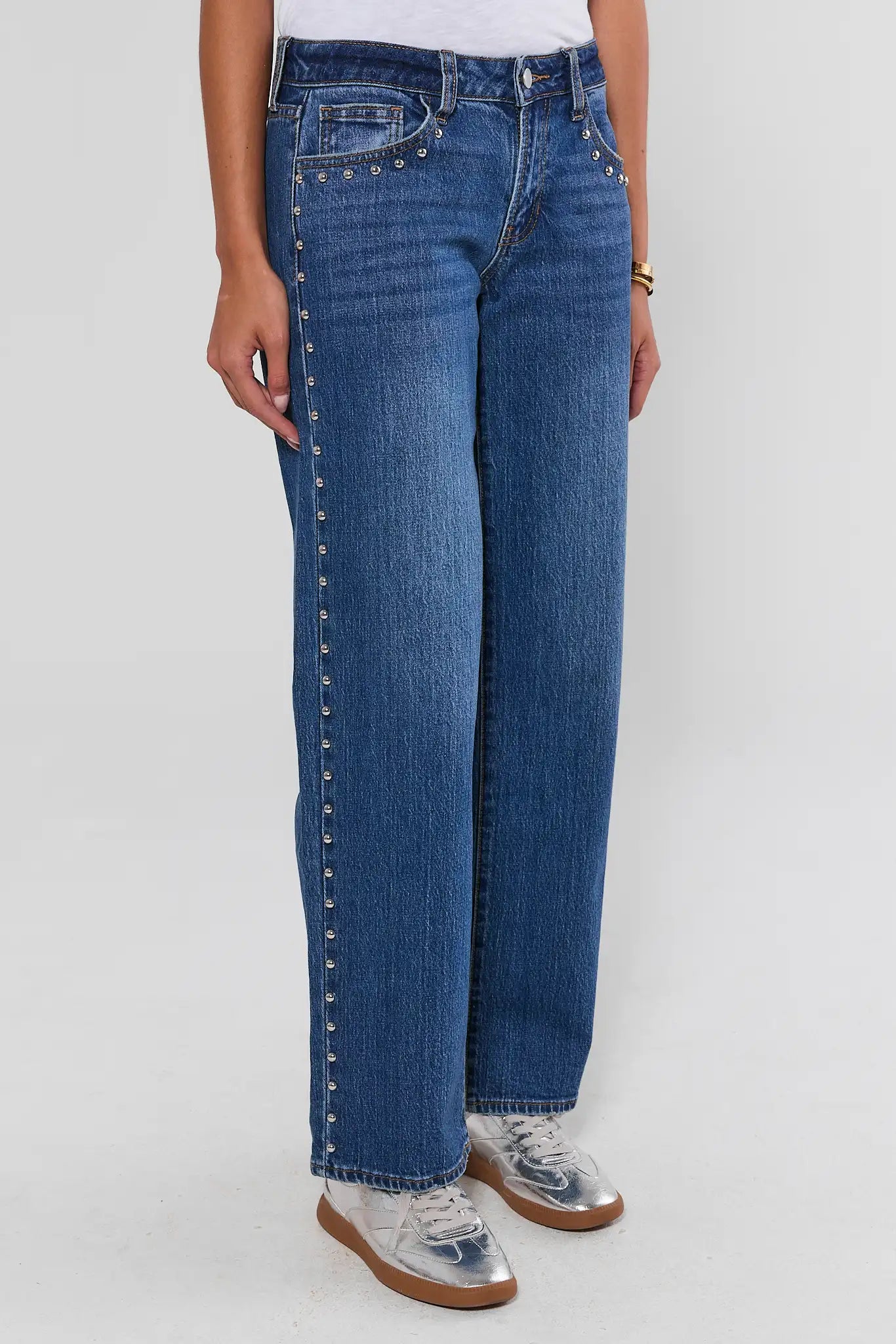 A person wearing blue denim jeans with silver stud detailing running down the outer seams, paired with a white top and metallic silver sneakers, shown against a light gray studio background.