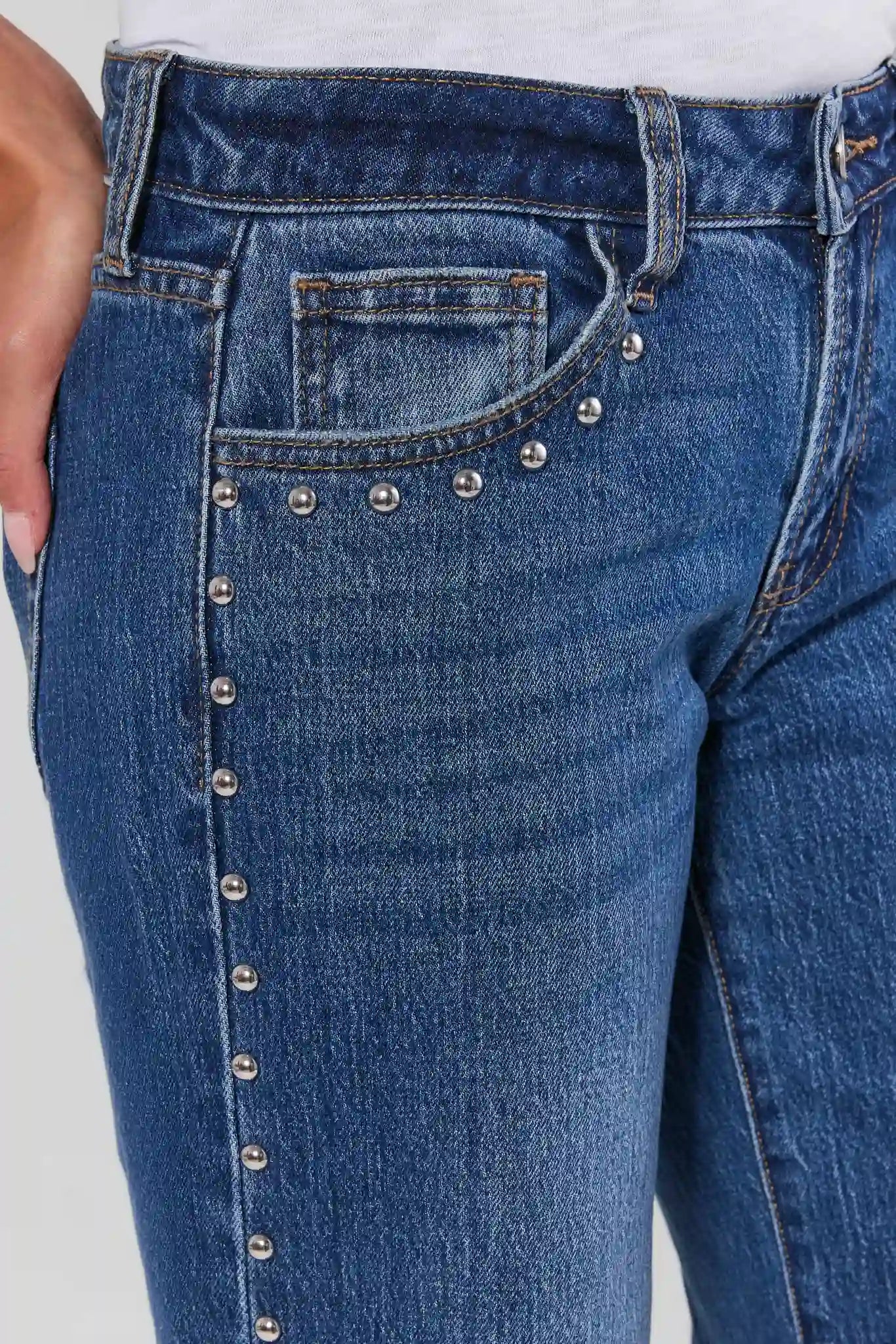 Close-up of blue denim jeans with decorative silver-tone studs running vertically along the front pocket and waistband area, worn with a white top.