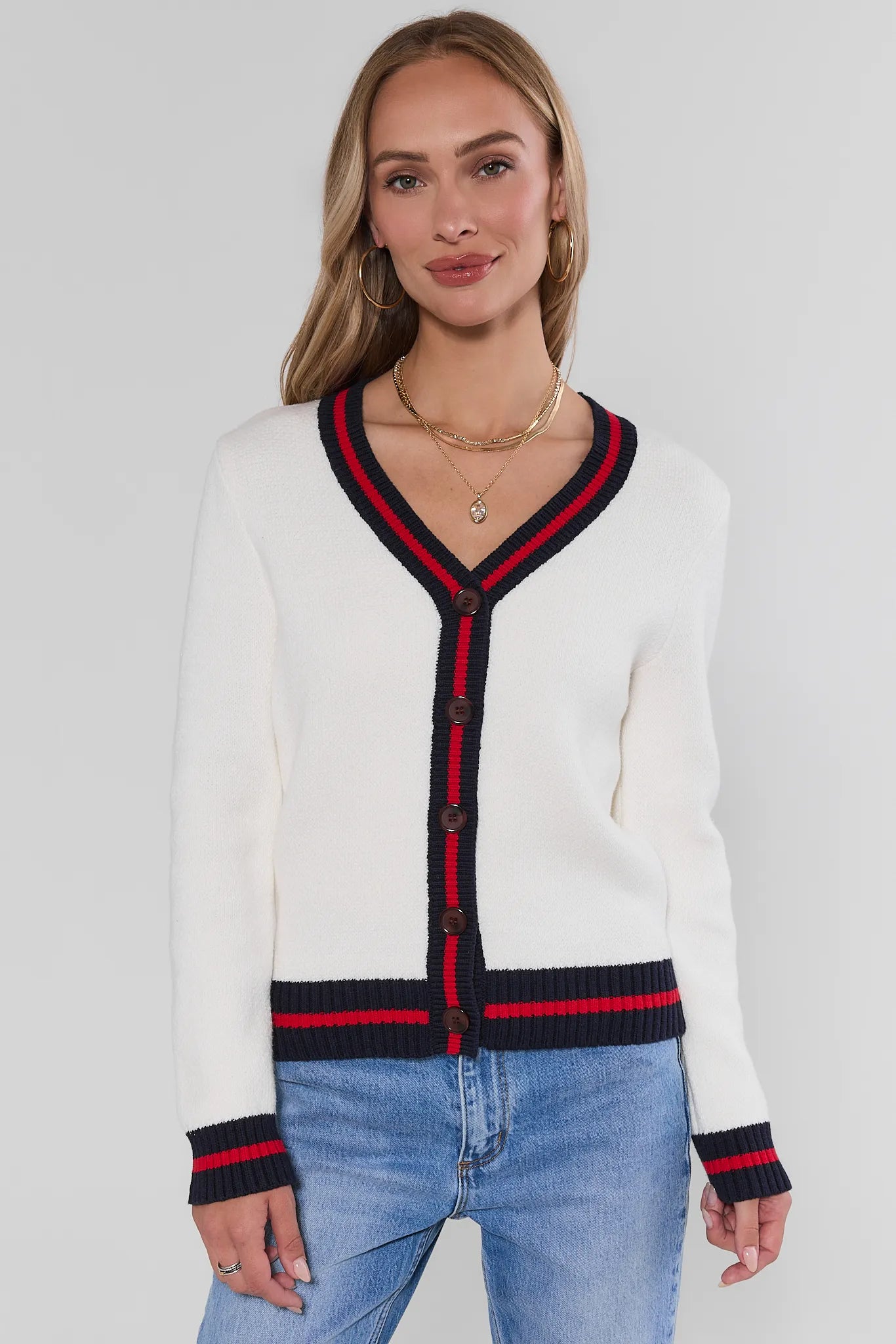 A woman wearing a white knit cardigan with navy and red striped trim on the V-neck, front buttons, cuffs, and hem, paired with light blue jeans against a light gray studio background.