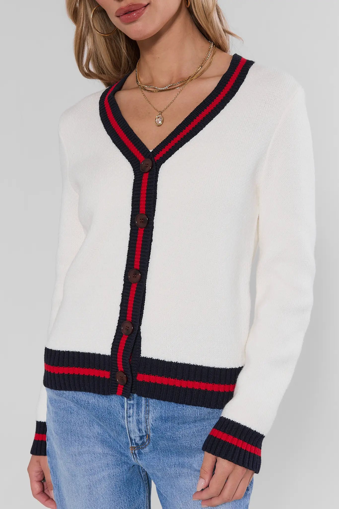 A woman wearing a white knit cardigan with navy and red striped trim along the V-neck, front buttons, hem, and cuffs, paired with light blue jeans against a light gray studio background.