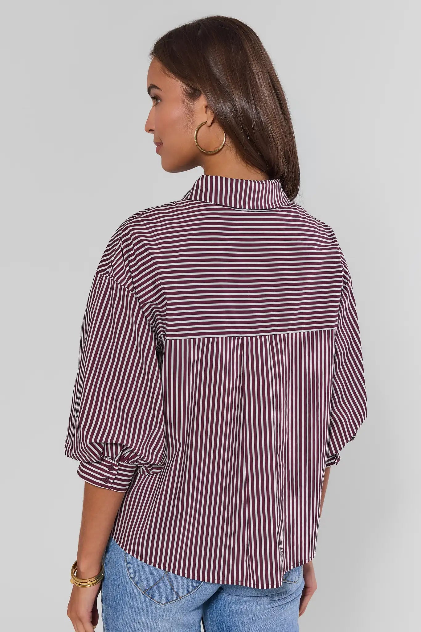 A woman shown from the back wearing a burgundy and white vertically striped blouse with balloon sleeves, paired with blue jeans, against a light gray studio background.
