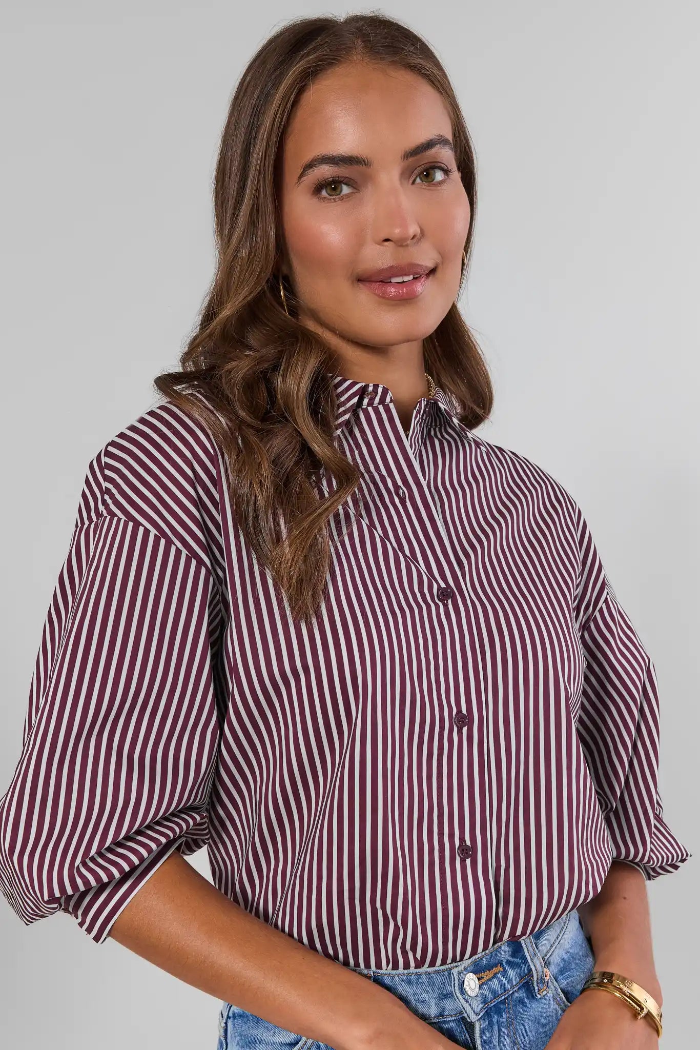A woman wearing a burgundy and white vertically striped button-up shirt with puff sleeves, paired with blue jeans, against a plain light gray background.