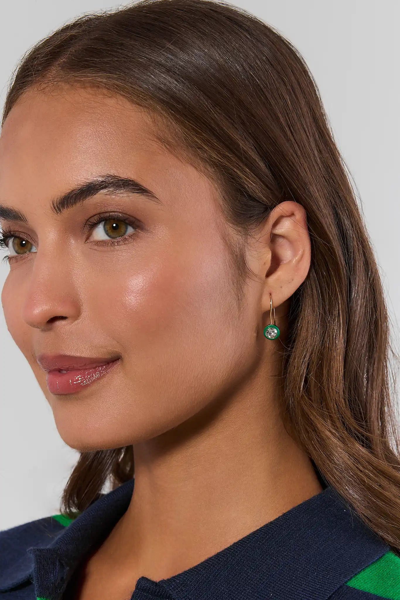 A close-up of a woman wearing a small gold hoop earring with a round green gemstone, shown against a light gray background.