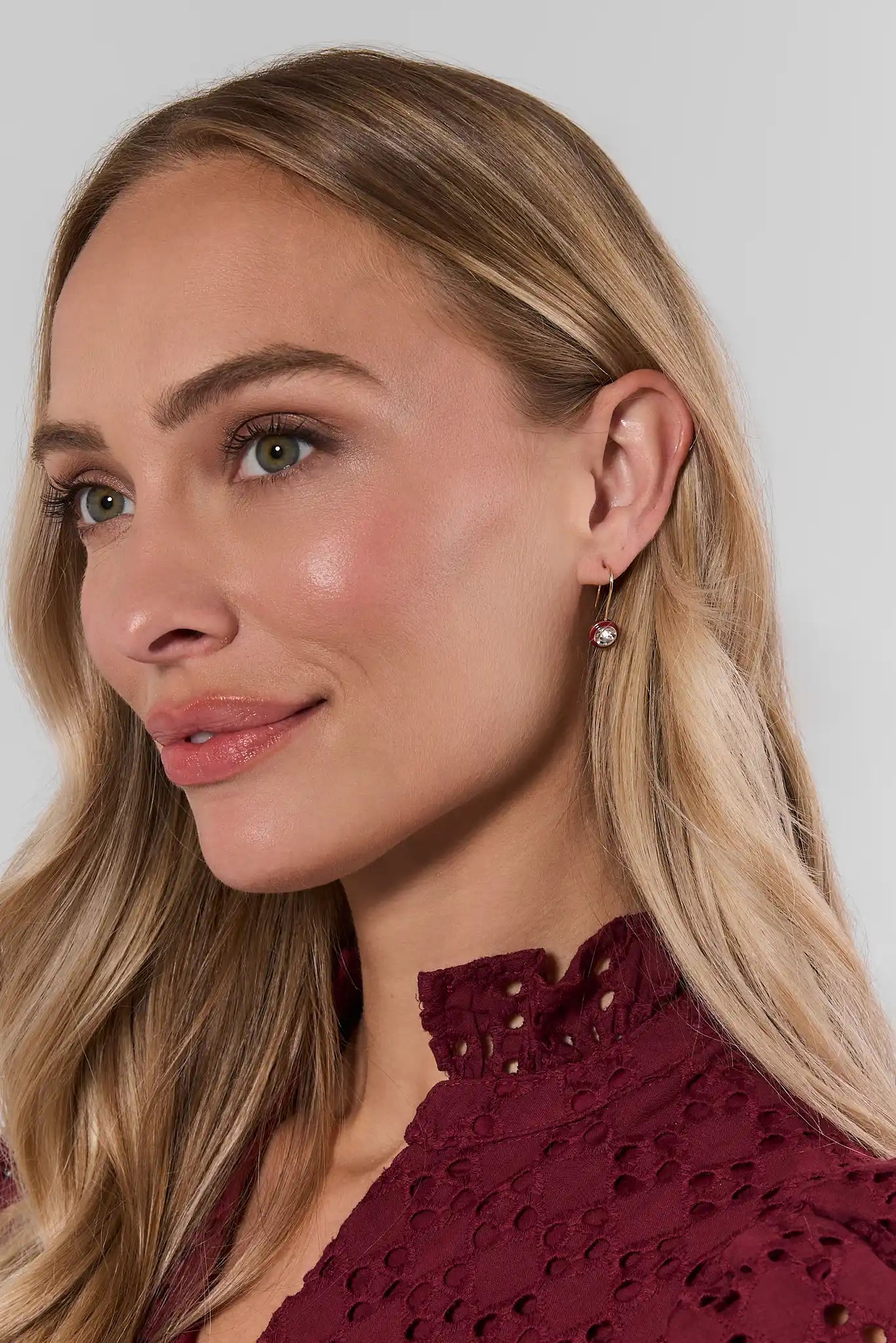 A close-up of a woman with blonde hair wearing a small gold hoop earring with a clear crystal pendant, paired with a burgundy eyelet top against a light gray background.