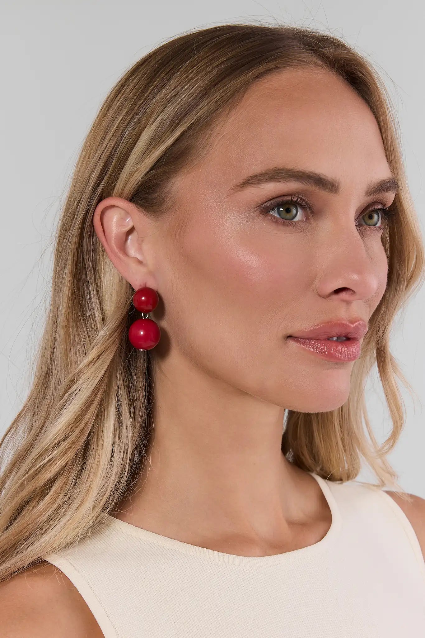 A close-up of a woman with blonde hair wearing a pair of chunky, double-tiered red beaded drop earrings against a light gray background.