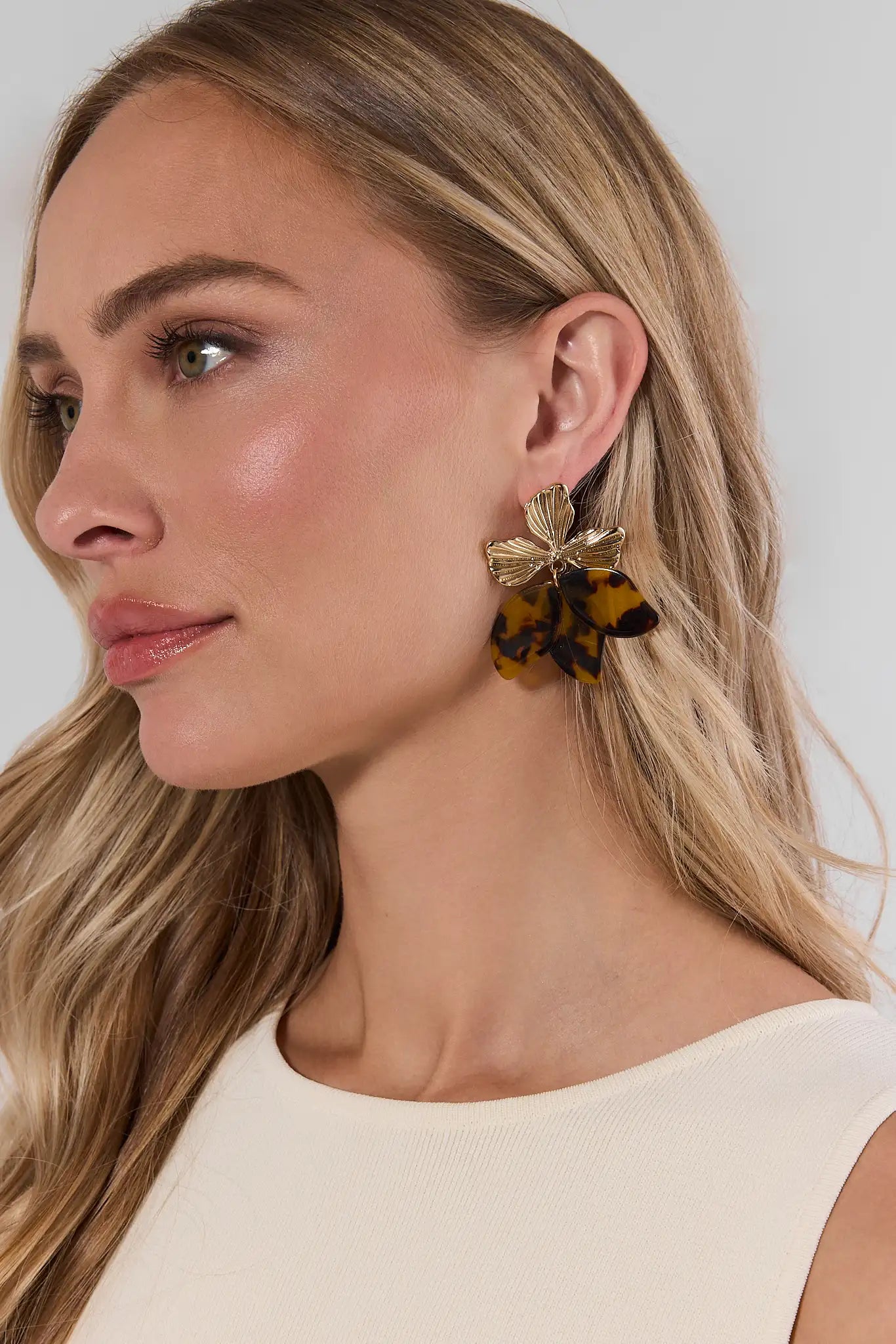 A close-up side profile of a woman with blonde hair wearing a dangling earring. The earring features a gold-toned metal top with layered translucent amber and dark brown petal-like drops, shown against a light neutral background.