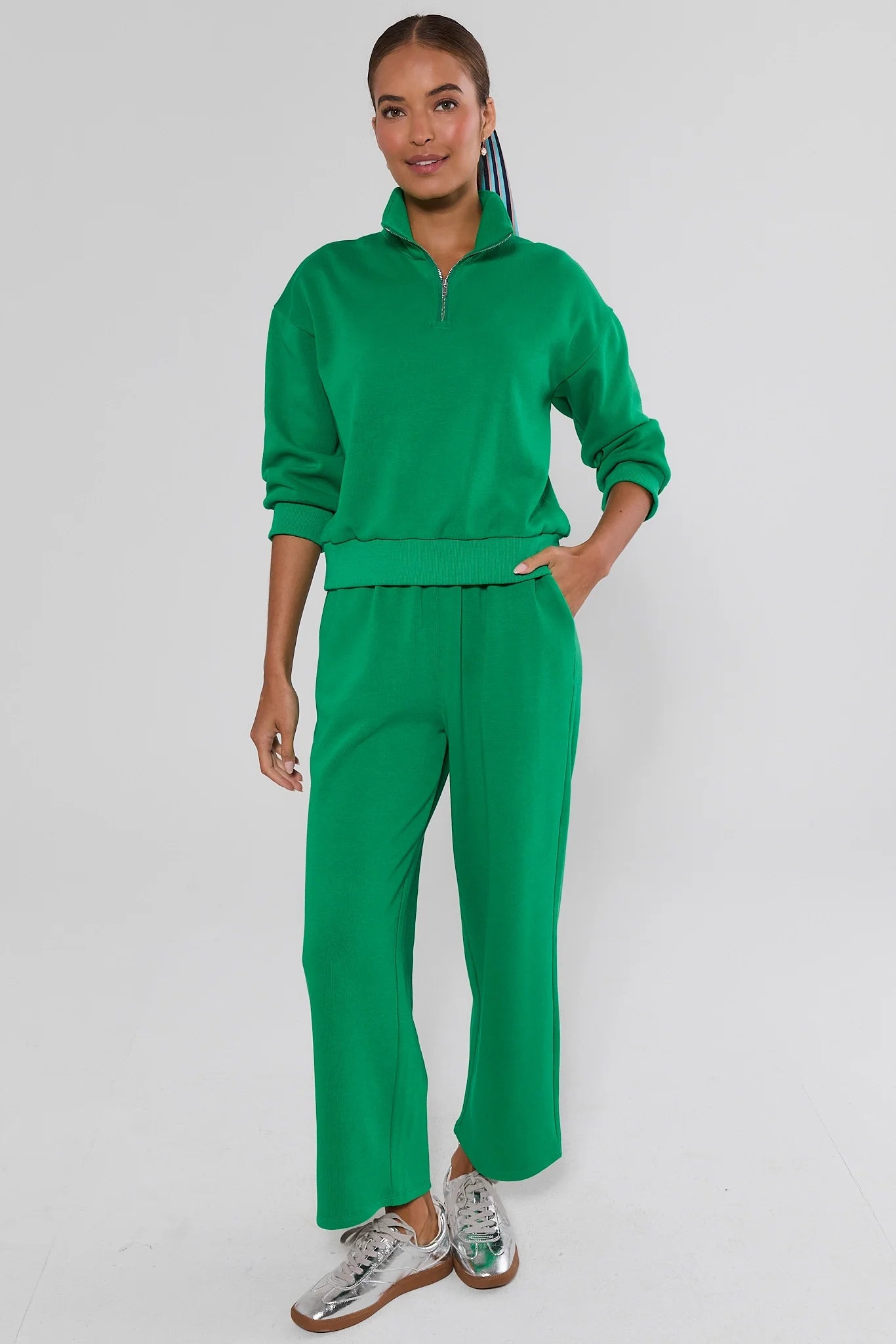 A woman modeling a matching bright green two-piece tracksuit with a half-zip long-sleeve top and wide-leg cropped pants, paired with silver sneakers, against a light background.