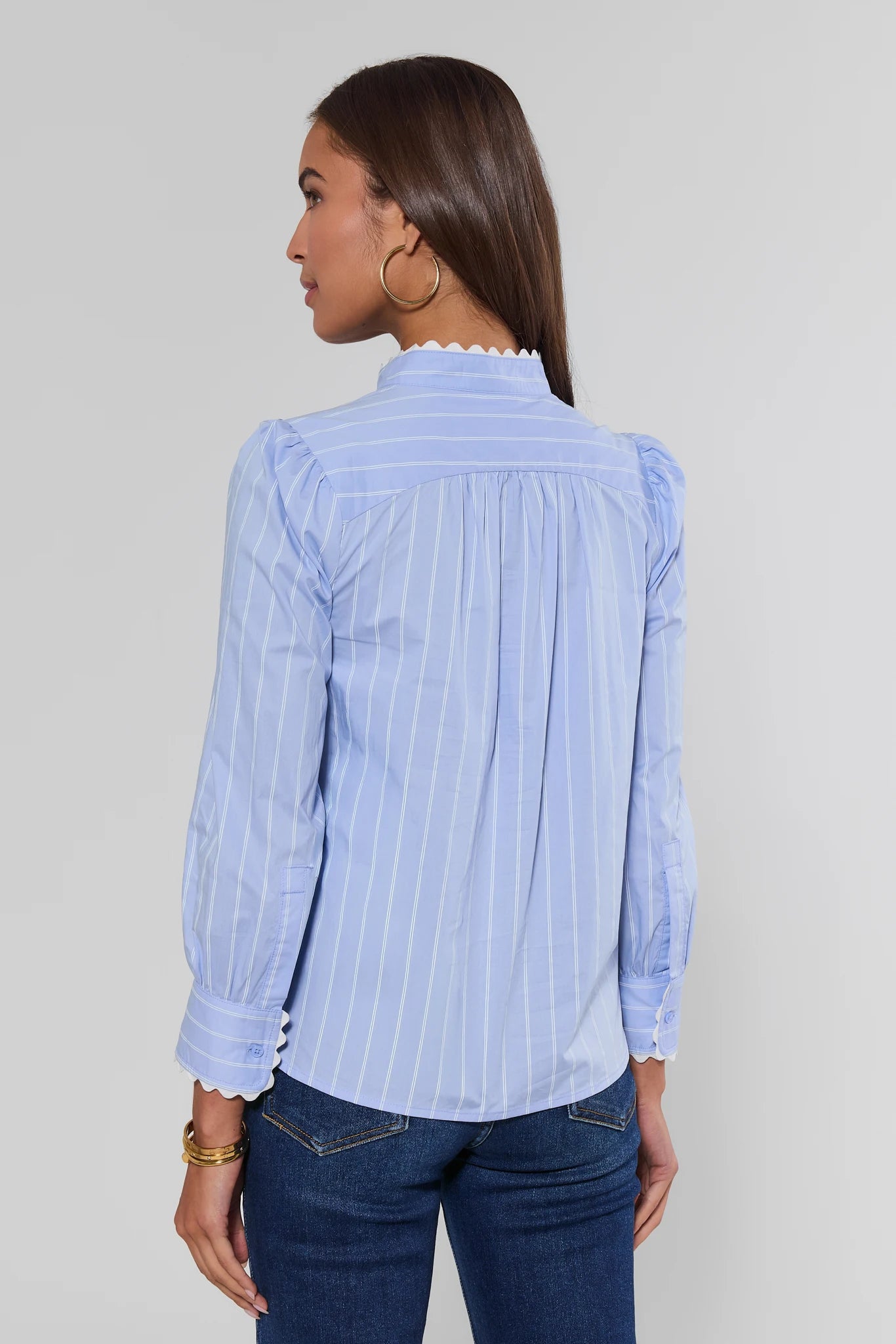 A woman shown from the back wearing a light blue and white vertically striped blouse with ruffled trim at the collar and cuffs, paired with dark blue jeans, against a plain light gray studio background.