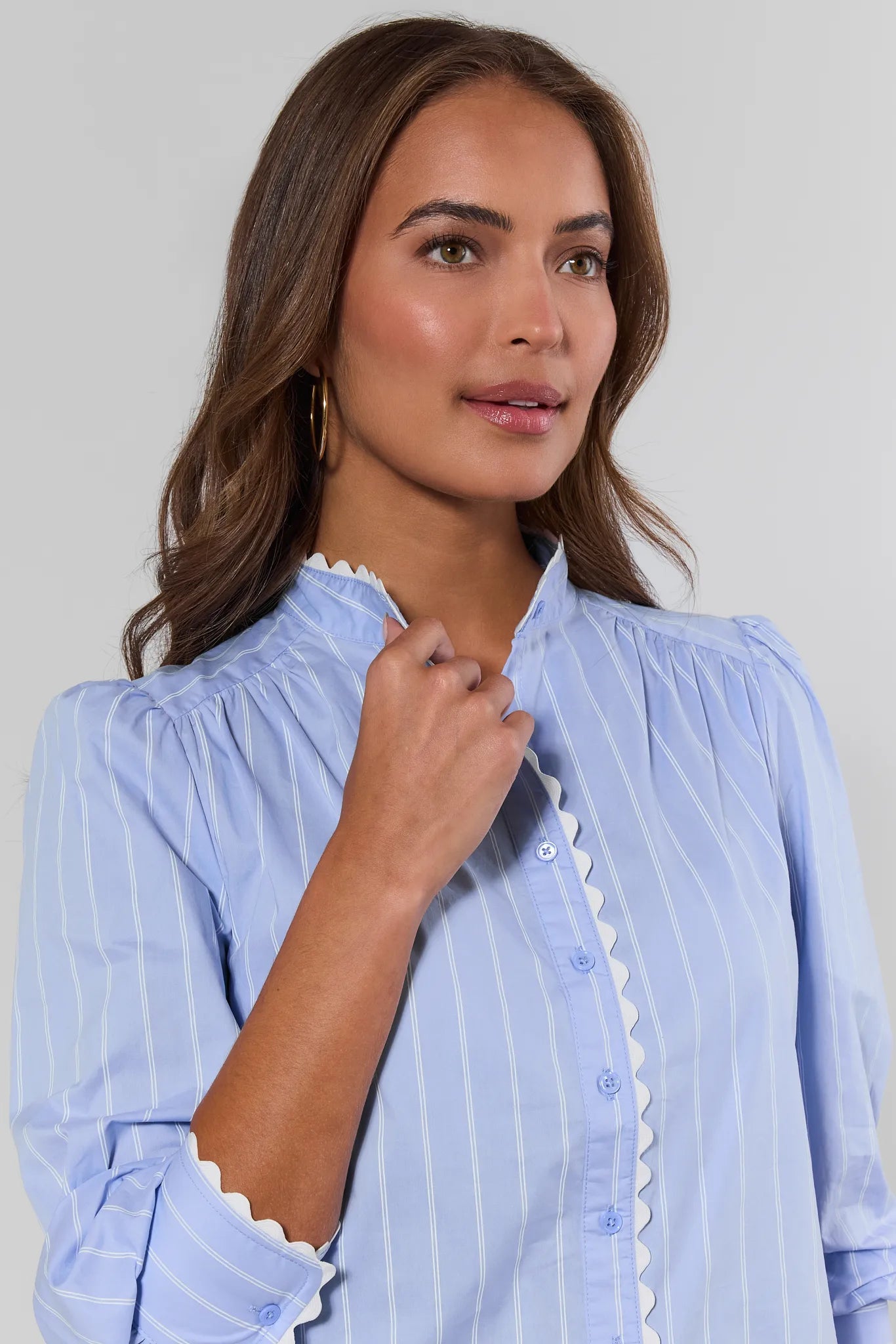 A woman wearing a light blue and white vertically striped blouse with white scalloped trim along the collar and front placket, shown against a plain light gray studio background.