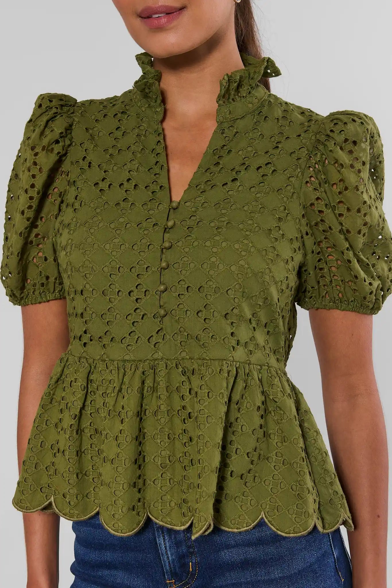 An olive green eyelet blouse with puff sleeves, a ruffled high neckline, and a scalloped peplum hem, shown on a model wearing blue jeans.