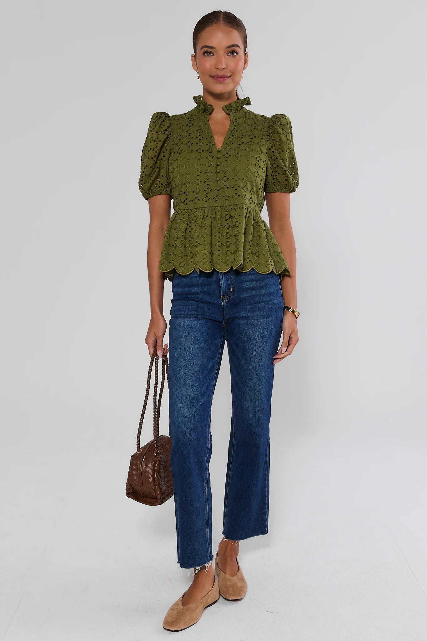 A woman modeling an olive green lace puff-sleeve blouse with a ruffled scalloped hem, paired with blue jeans, tan flats, and a brown handbag against a light gray studio background.