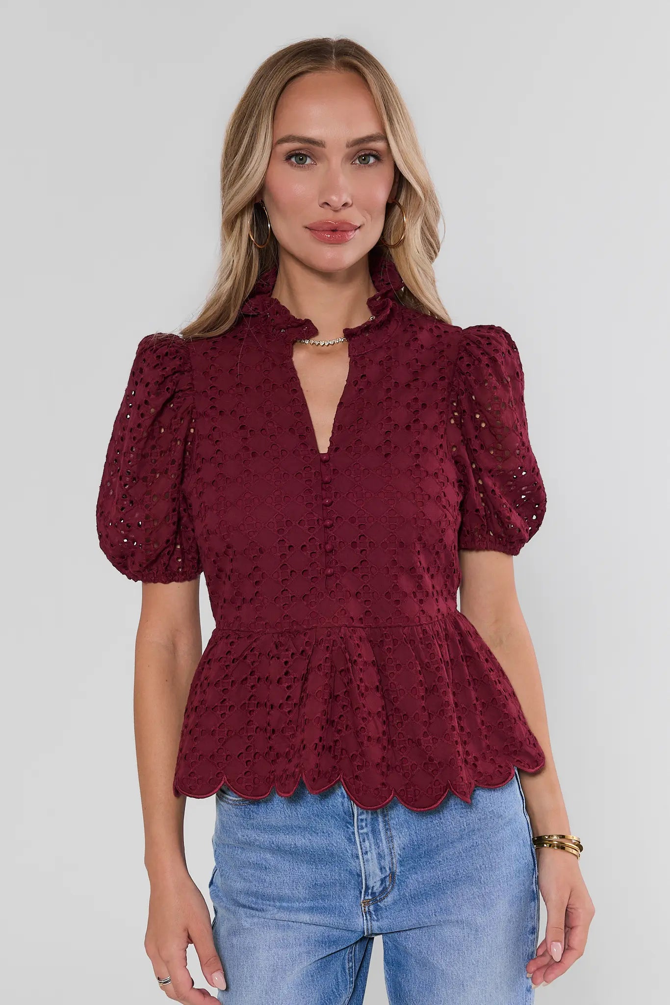 A woman wearing a burgundy eyelet lace top with puff sleeves and a scalloped peplum hem, paired with blue jeans, against a light gray studio background.