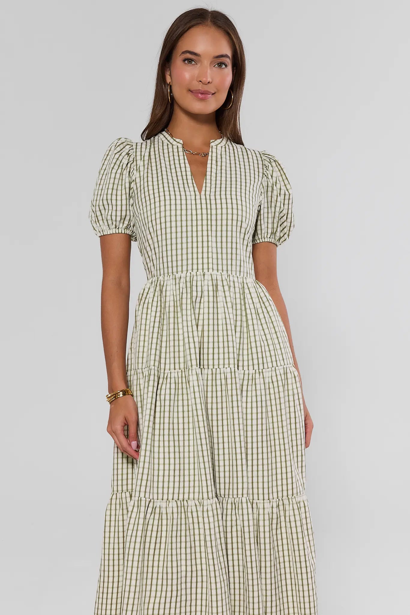 A woman modeling a tiered midi dress with puff sleeves and a green and white gingham pattern, set against a plain light gray studio background.