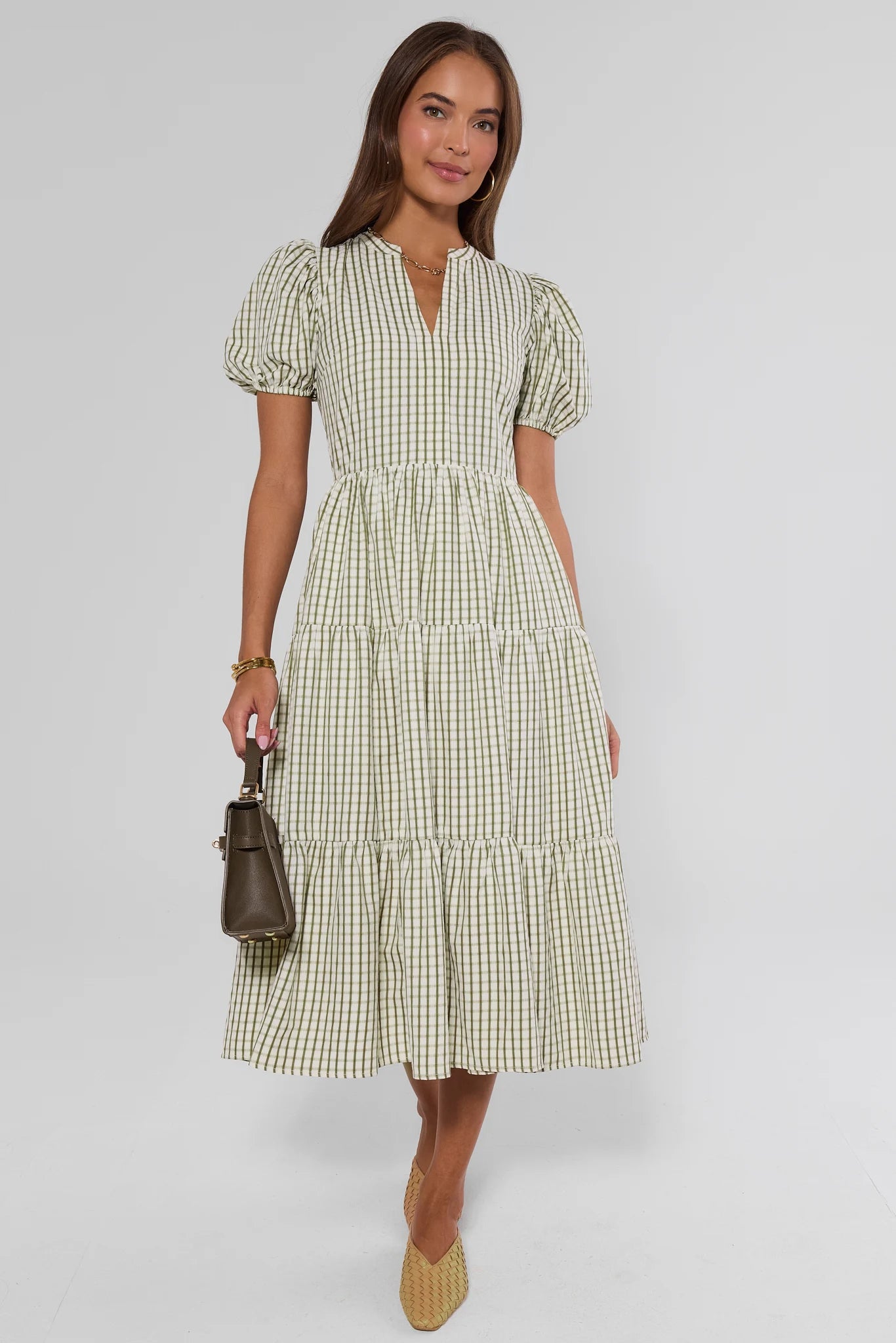 A woman modeling a light green and white gingham midi dress with puff sleeves, a split neckline, and a tiered skirt, holding a small brown handbag against a light gray studio background.