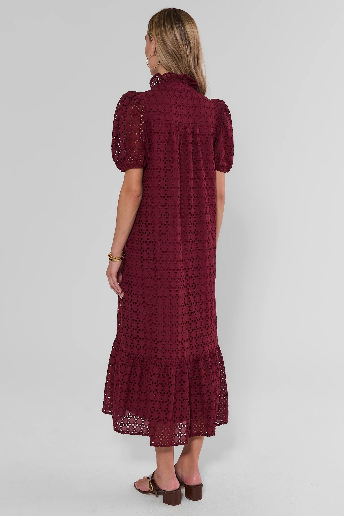 A woman viewed from the back wearing a dark burgundy eyelet midi dress with puff sleeves and a ruffled hem, paired with brown sandals, against a light gray studio background.