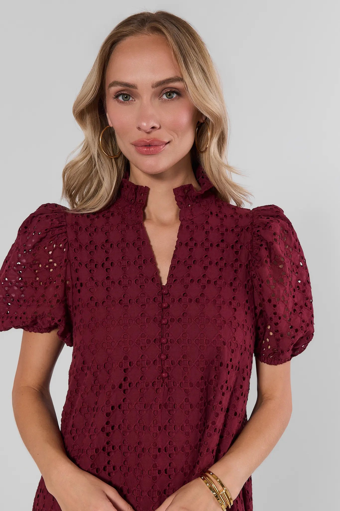 A woman wearing a burgundy eyelet lace top with puff sleeves and a split neckline, photographed against a light gray studio background.