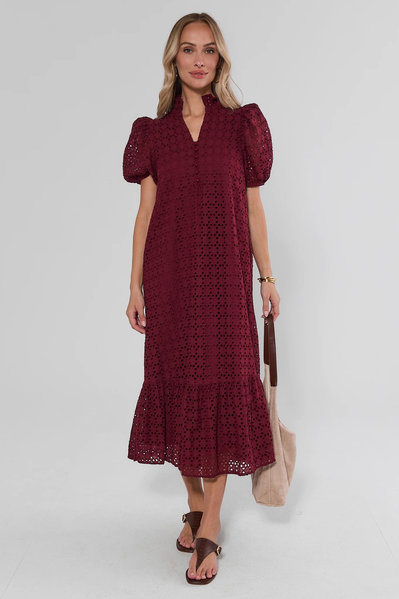 A woman modeling a deep burgundy midi dress with puff sleeves, a V-neckline, and an intricate eyelet lace pattern, standing against a light gray studio background.