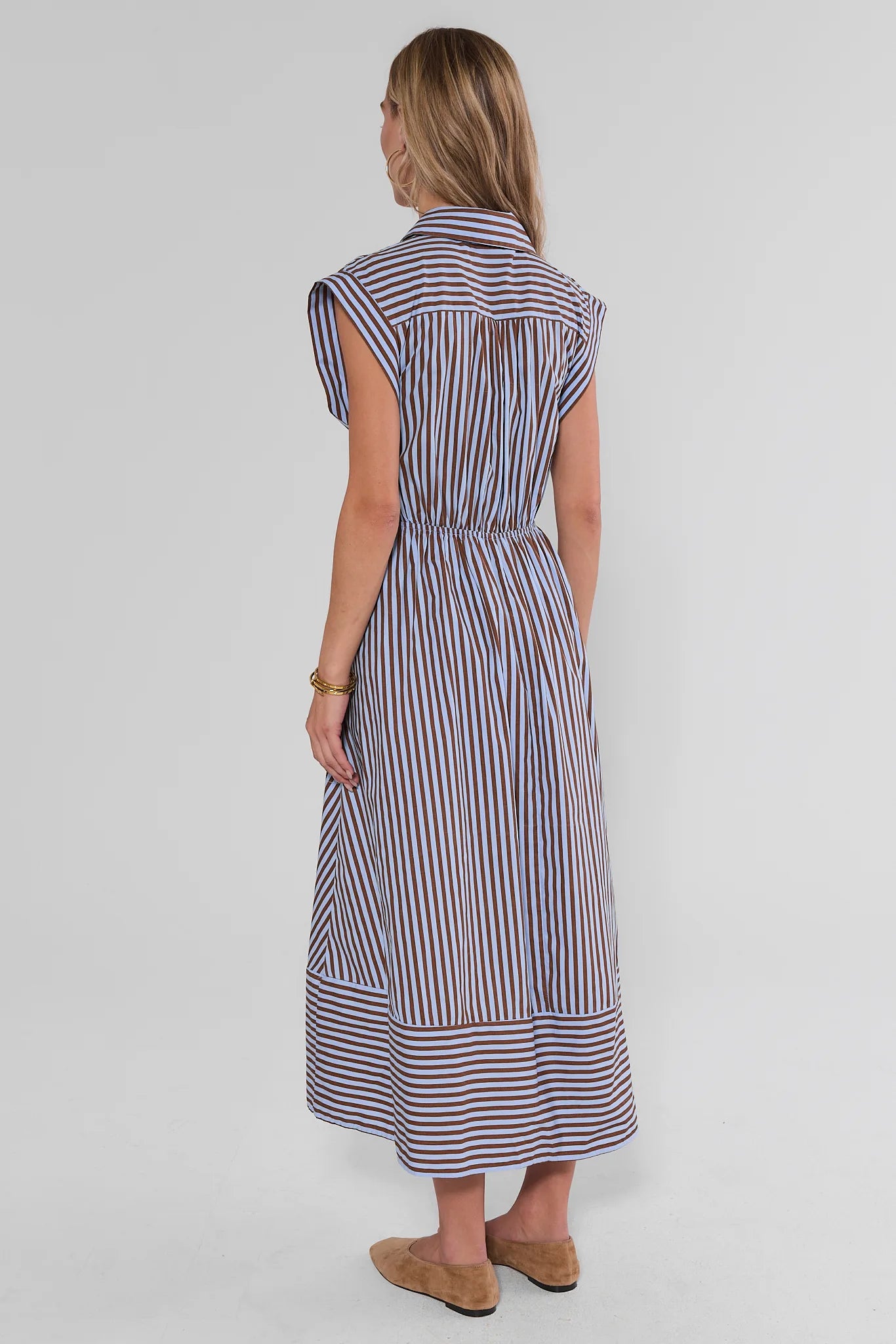 A woman shown from the back wearing a sleeveless midi dress with vertical blue, red, and white stripes, paired with tan flat shoes, against a light gray studio background.
