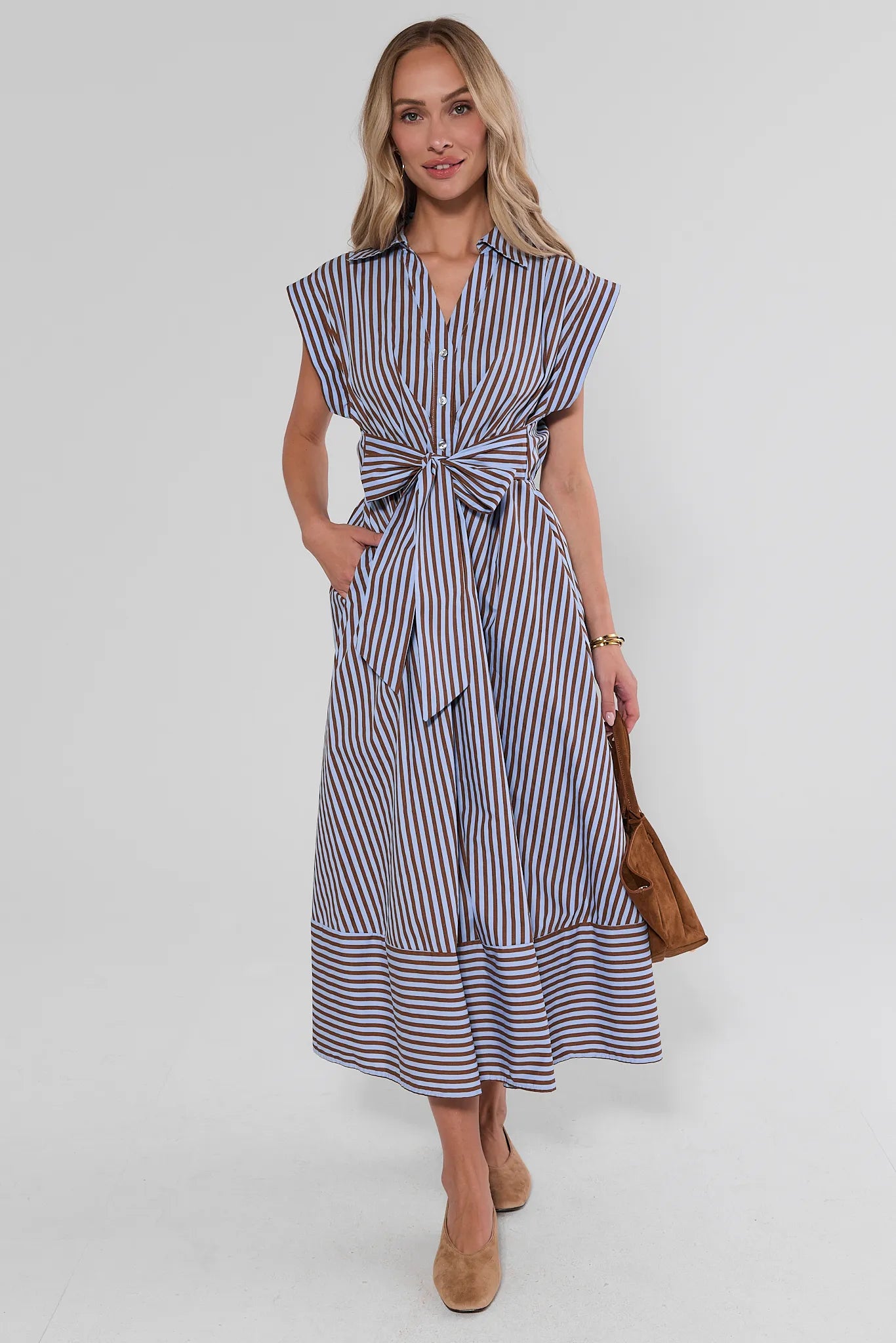 A woman modeling a sleeveless midi dress with vertical navy and white stripes, featuring a tie waist and ruffled hem, paired with beige heels and a brown handbag against a light gray studio background.