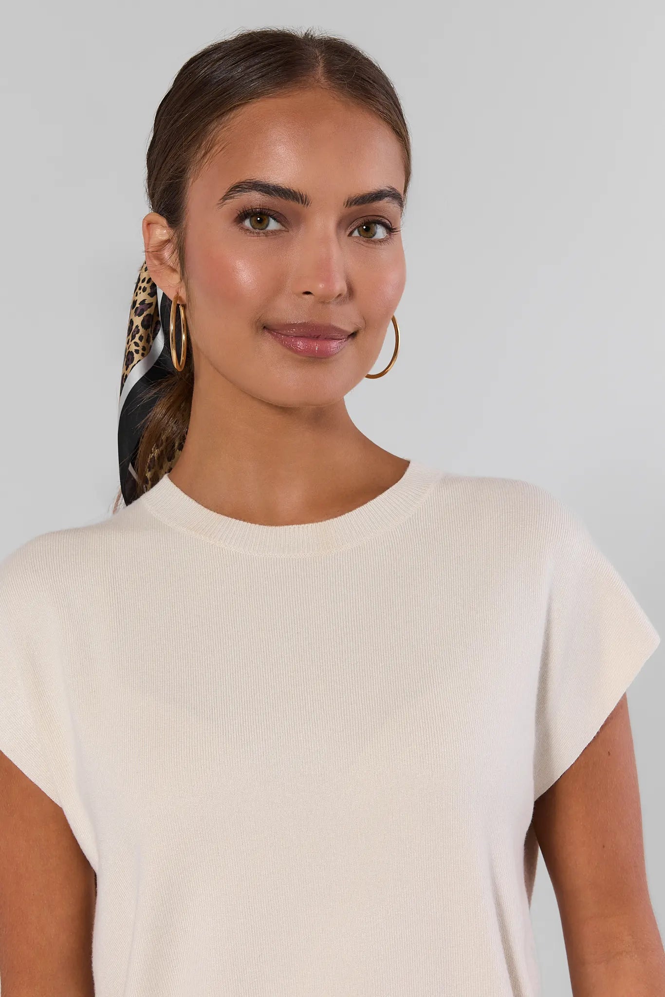A woman wearing a cream-colored short-sleeve crewneck top, styled with a black and gold patterned hair scarf and gold hoop earrings, against a light gray background.