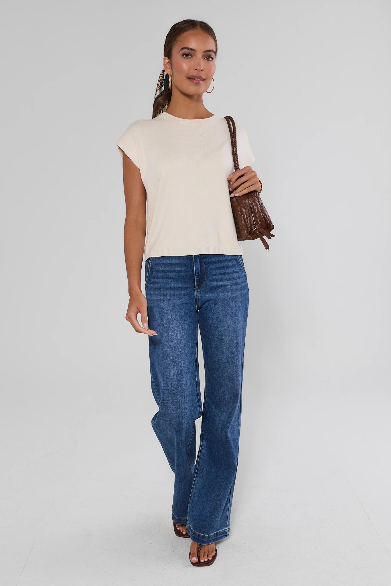 A woman modeling a cream short-sleeve top and blue wide-leg jeans, carrying a brown textured shoulder bag against a light studio background.