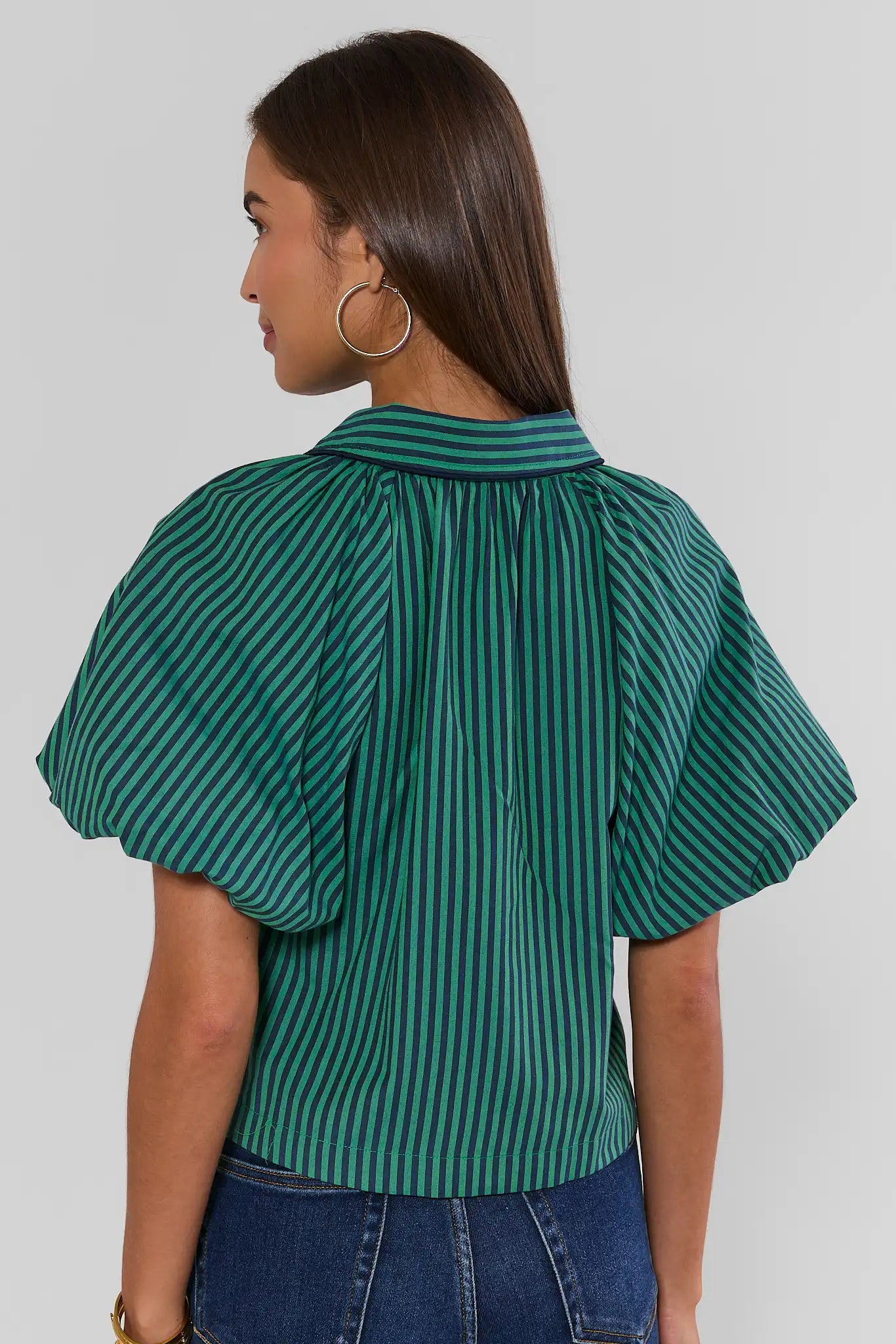 A woman shown from the back wearing a green and black vertically striped short-sleeve blouse with puffed sleeves, paired with blue jeans, against a light gray studio background.