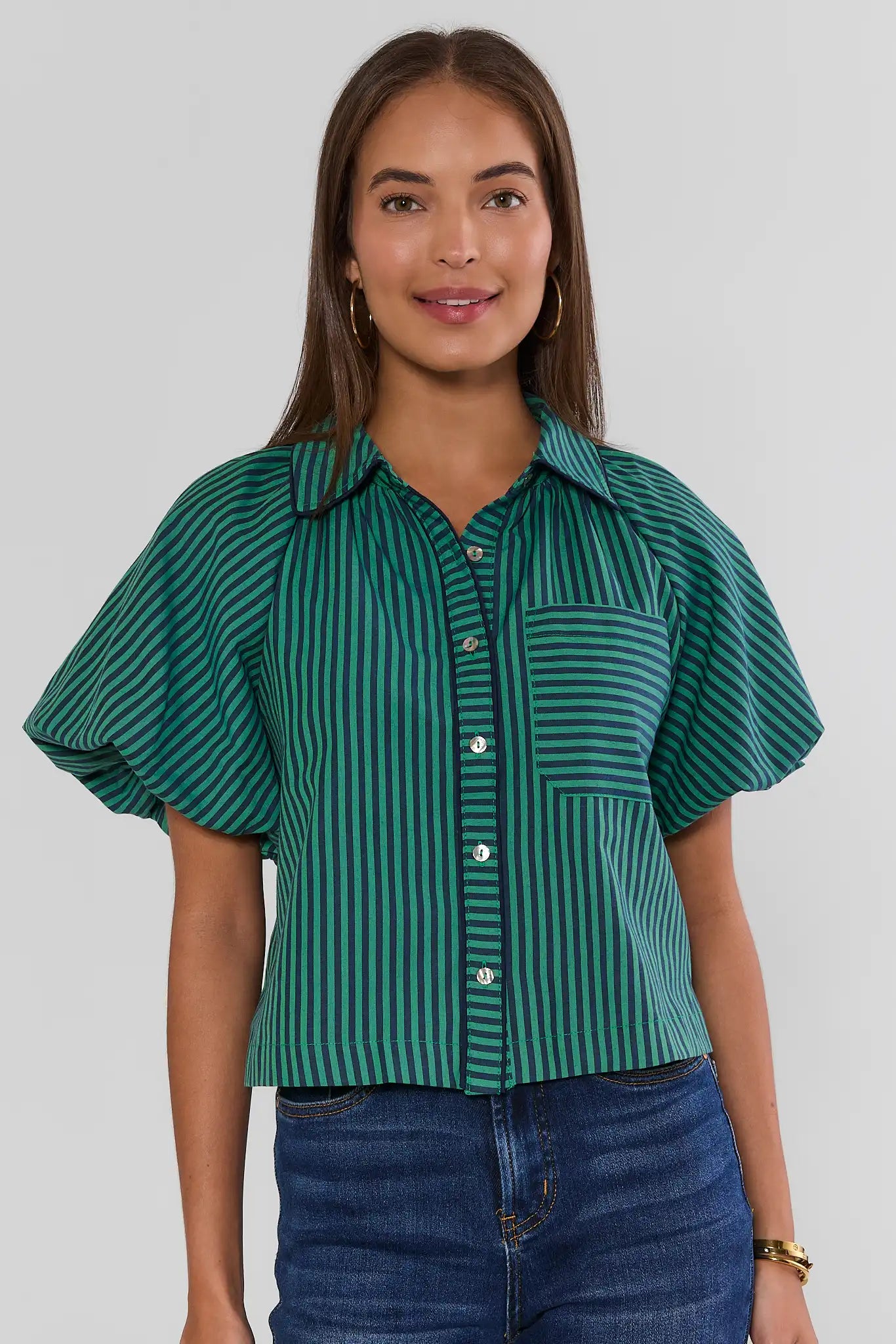 A woman wearing a green and black vertically striped short-sleeve button-up shirt with puff sleeves and a chest pocket, paired with blue jeans, against a light gray studio background.