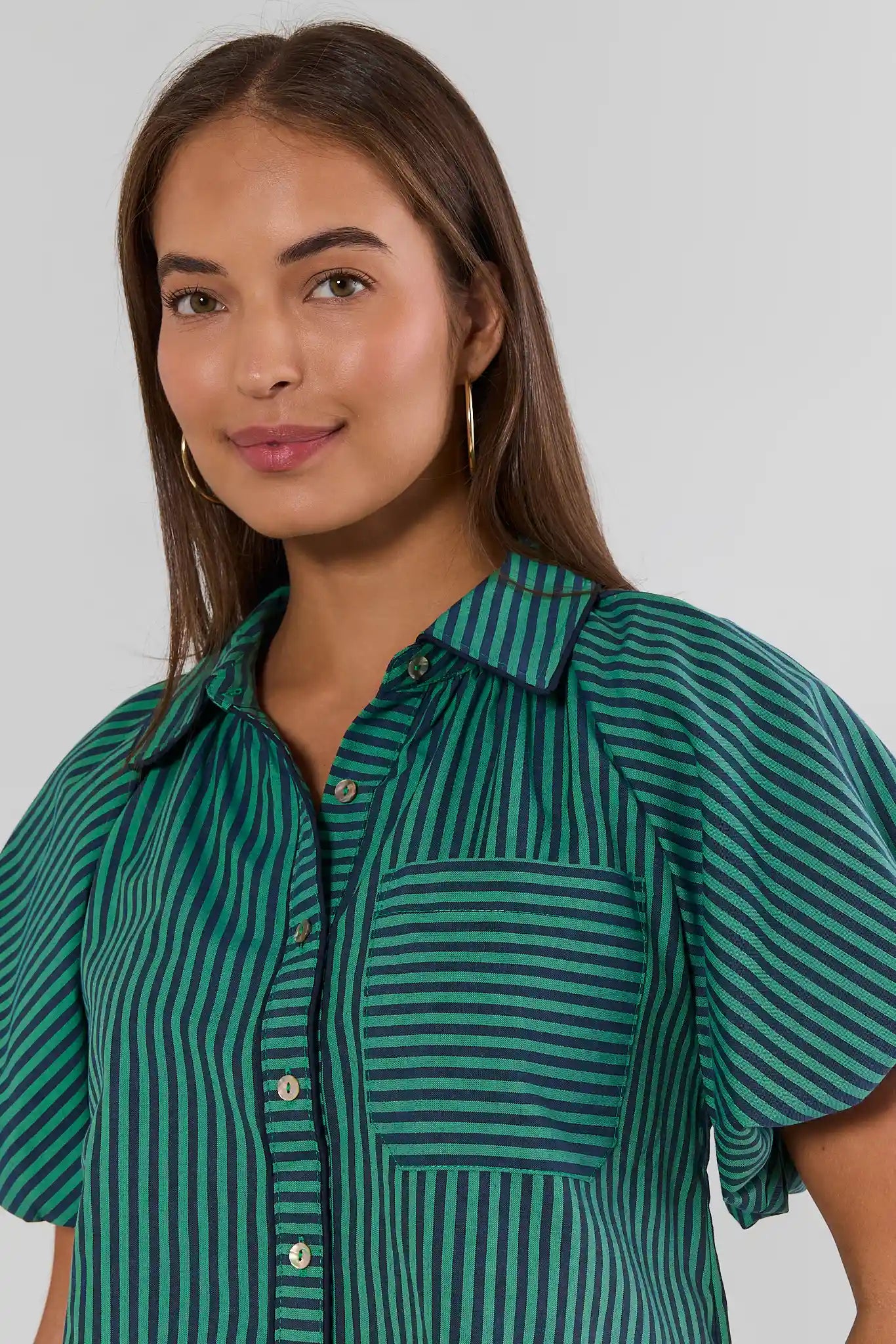 A woman wearing a green and dark teal striped short-sleeve button-up shirt, shown from the chest up against a light gray studio background.