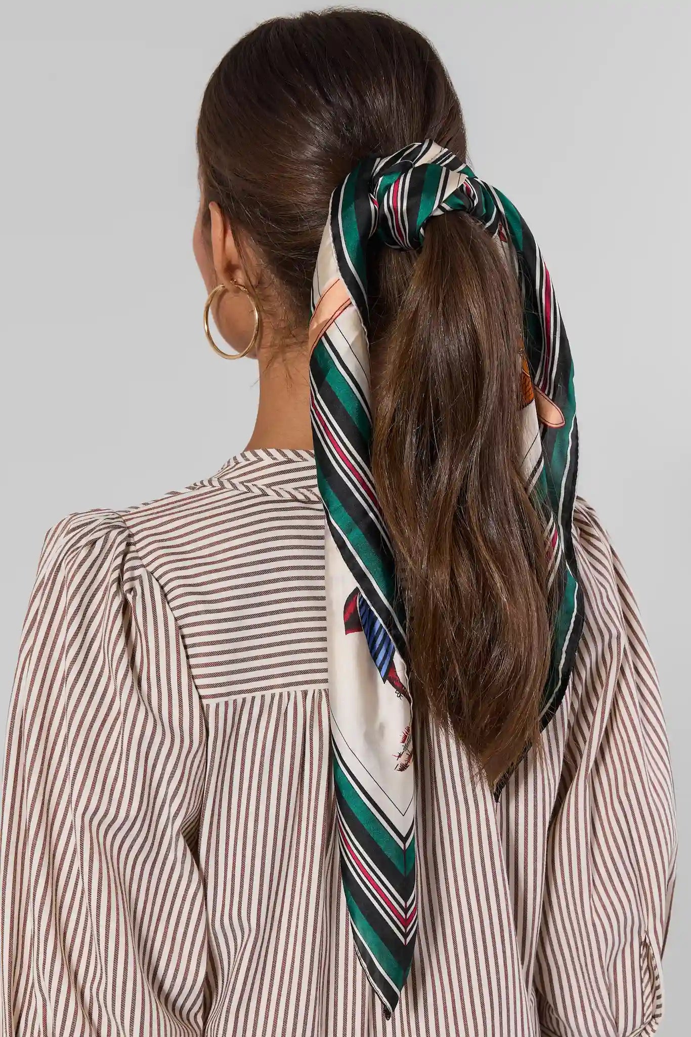 A woman shown from the back wearing a brown hair tie made from a patterned silk scarf with green, white, and red stripes, paired with a brown and white striped blouse.