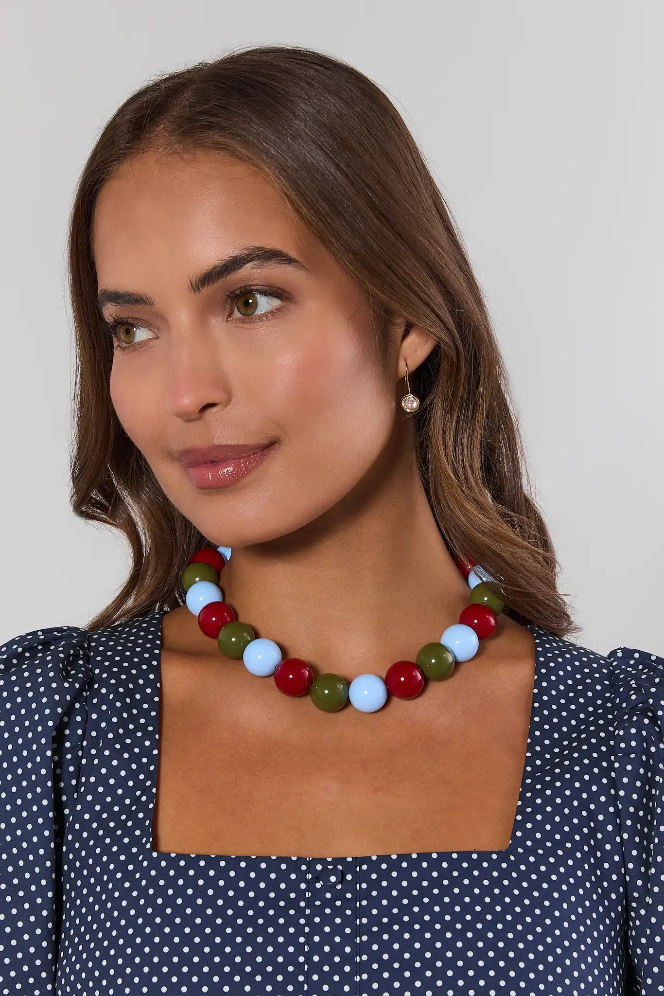 A woman wearing a chunky beaded necklace with red, light blue, olive green, and white beads, paired with a navy blue and white polka dot top against a light gray background.