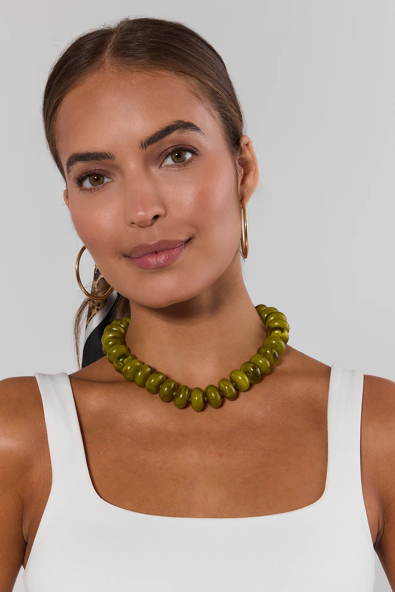 A woman wearing a chunky olive green beaded necklace and gold hoop earrings, shown from the chest up against a light gray studio background.