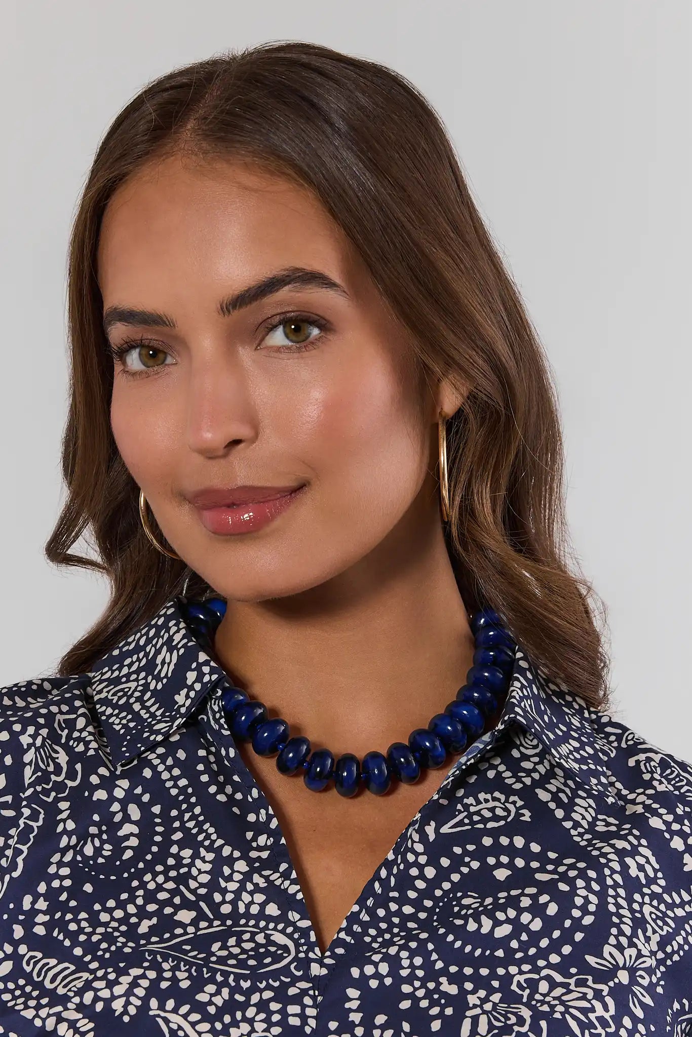 A woman with brown hair is wearing a dark blue beaded necklace and matching blue patterned blouse against a light gray background.