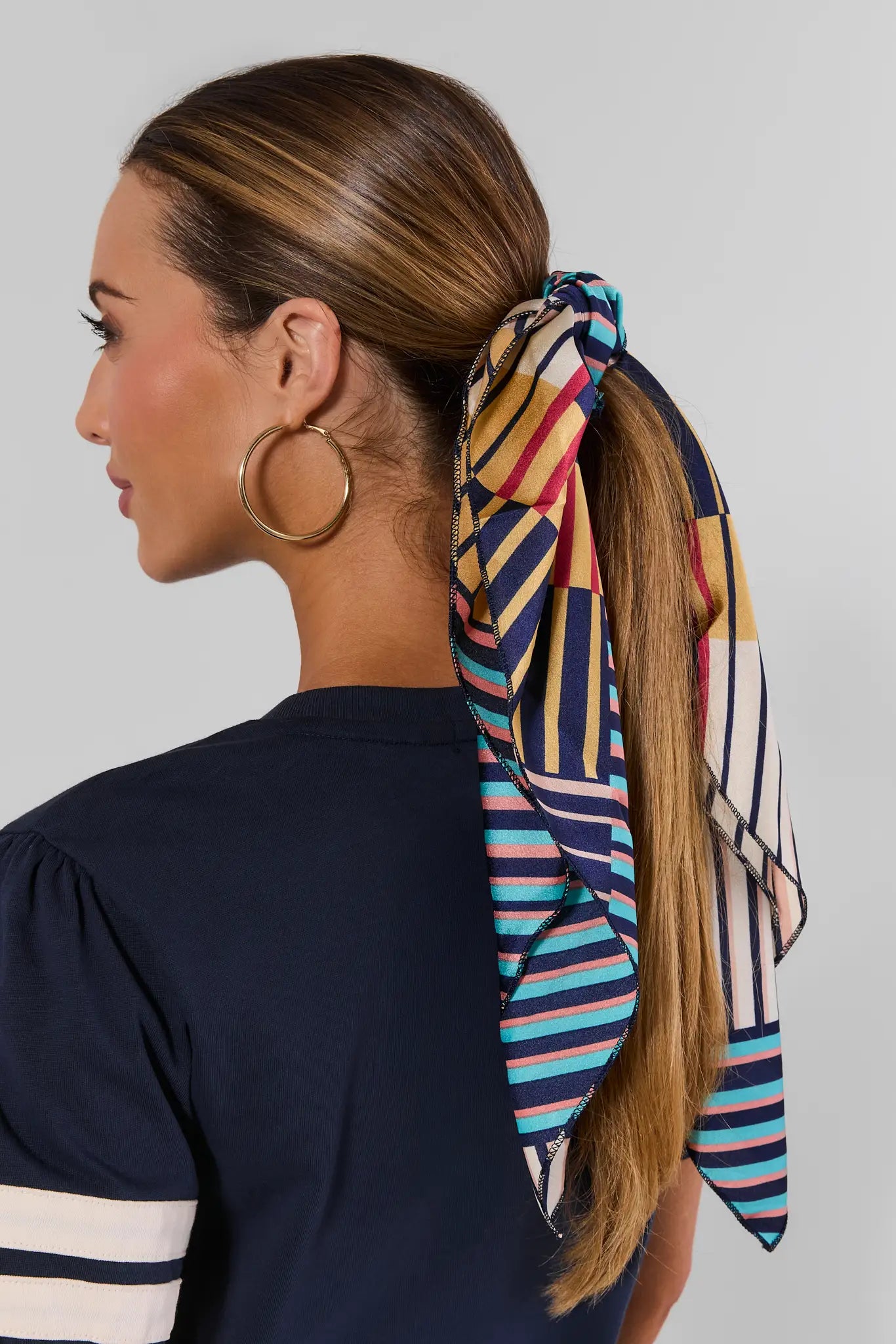 A woman shown from the back wearing a navy blue top and large gold hoop earrings, with her hair tied back in a ponytail using a multicolor striped scarf. The scarf features vertical stripes in navy, yellow, pink, and white.