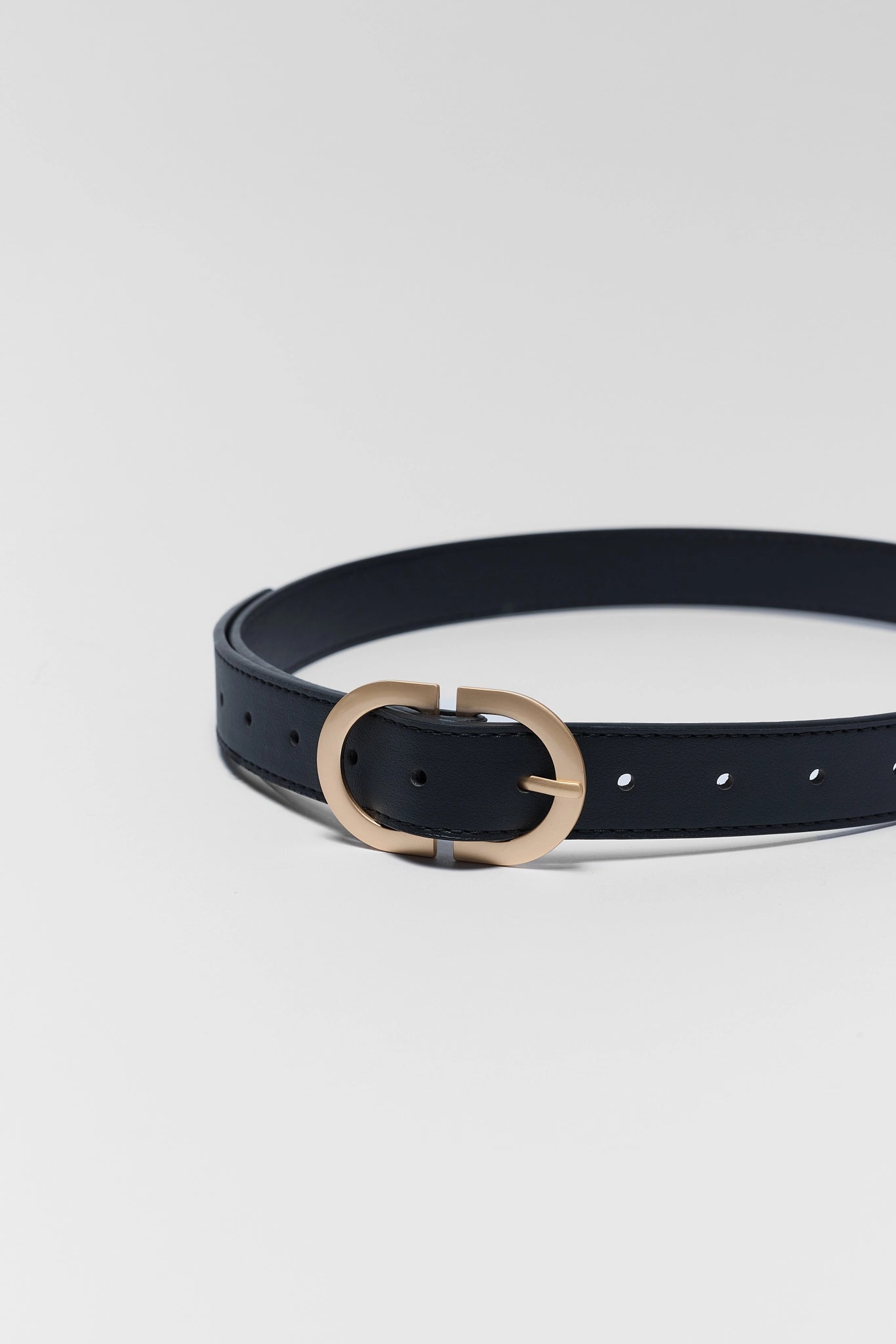 A slim black leather belt with a polished gold-tone oval buckle, coiled on a light gray studio background.