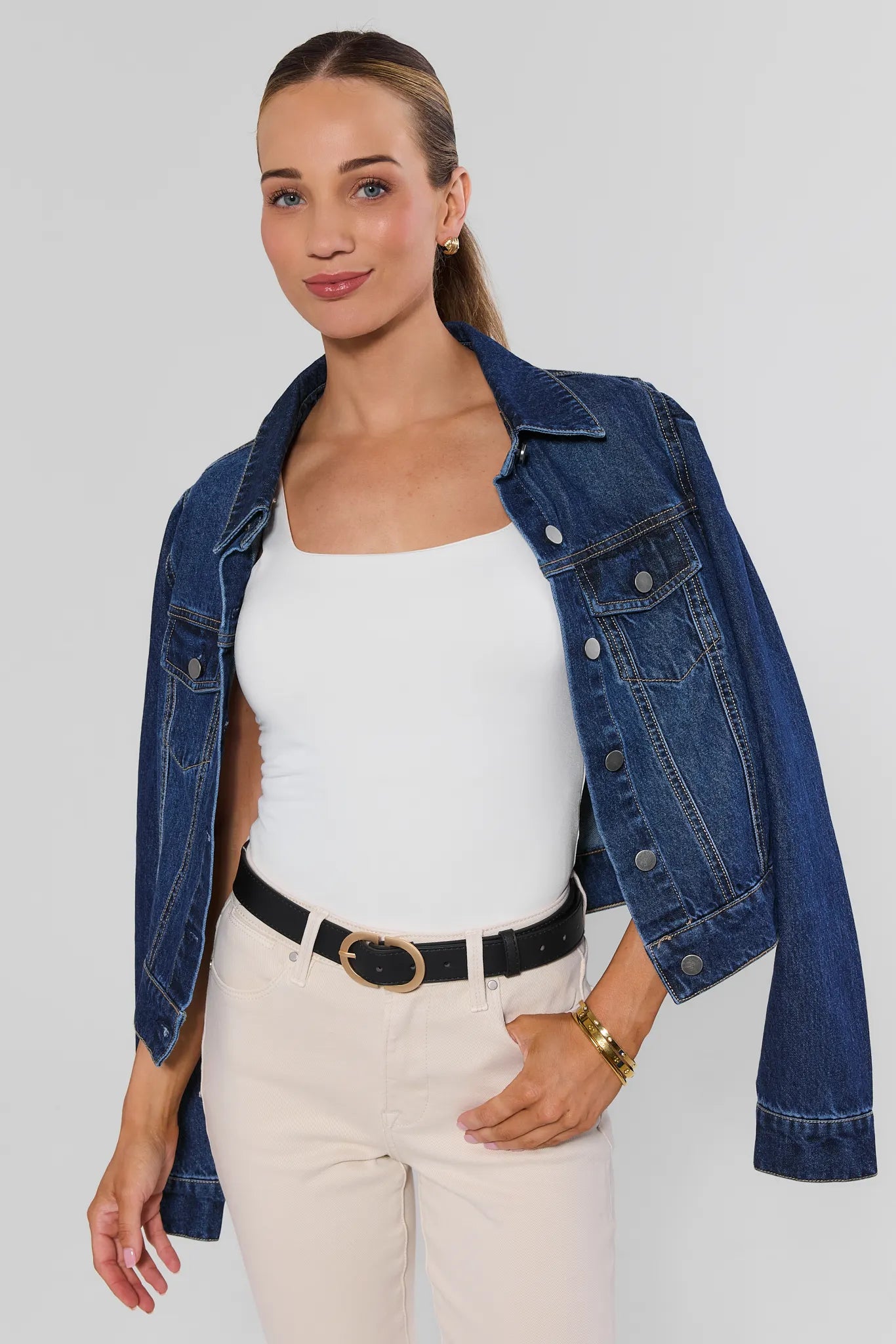 A woman modeling a cropped blue denim jacket over a white tank top and cream pants with a black belt, photographed against a light gray studio background.