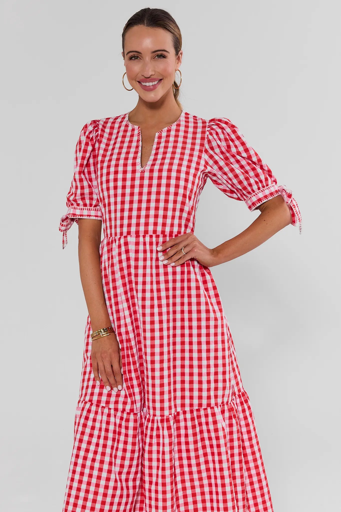 A woman modeling a red and white gingham tiered maxi dress with puff sleeves and a split neckline, photographed against a light gray studio background.
