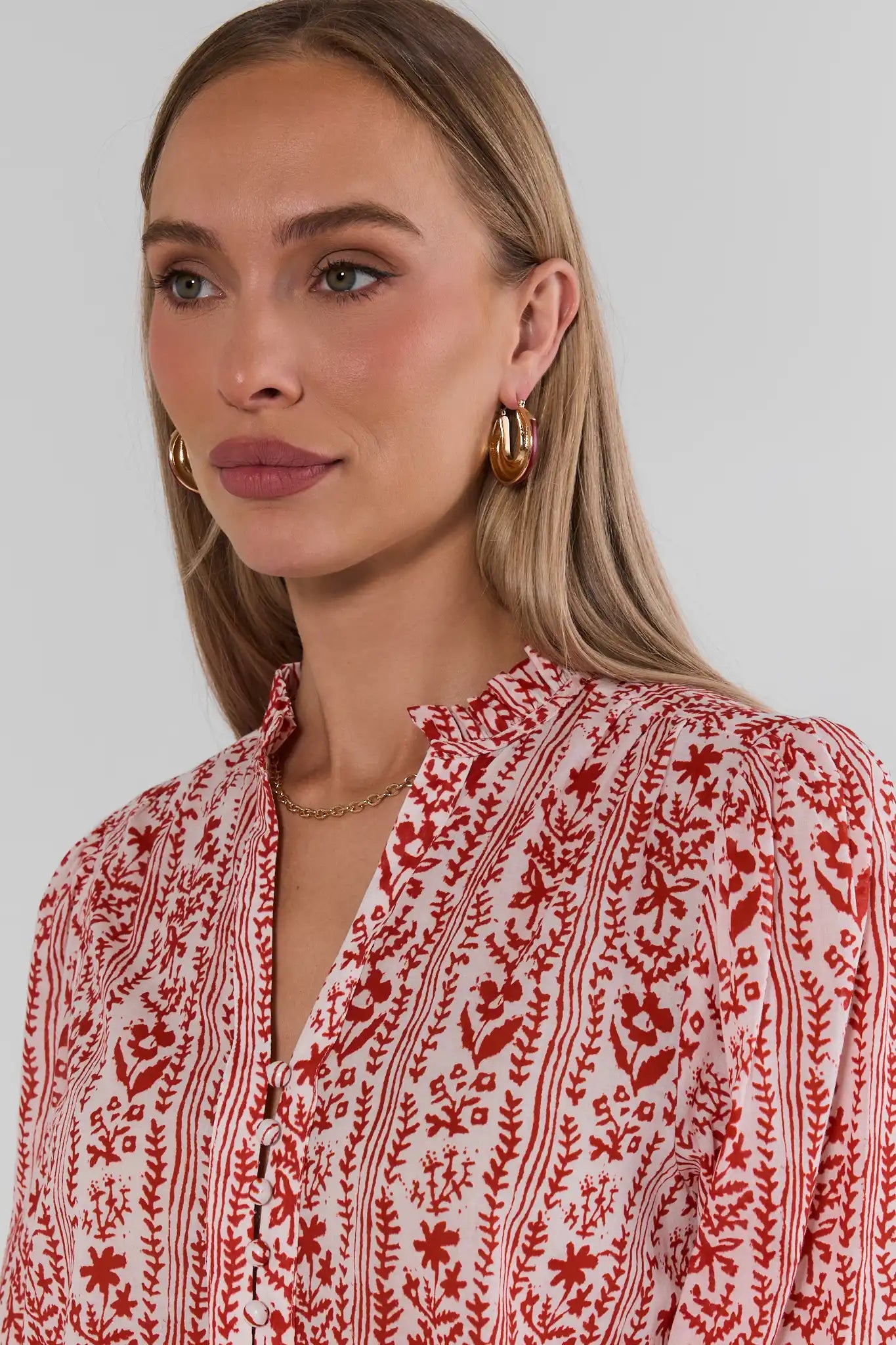 A close-up of a woman wearing Red Floral Printed Button Up Top with gold hoop earrings against a light gray background.