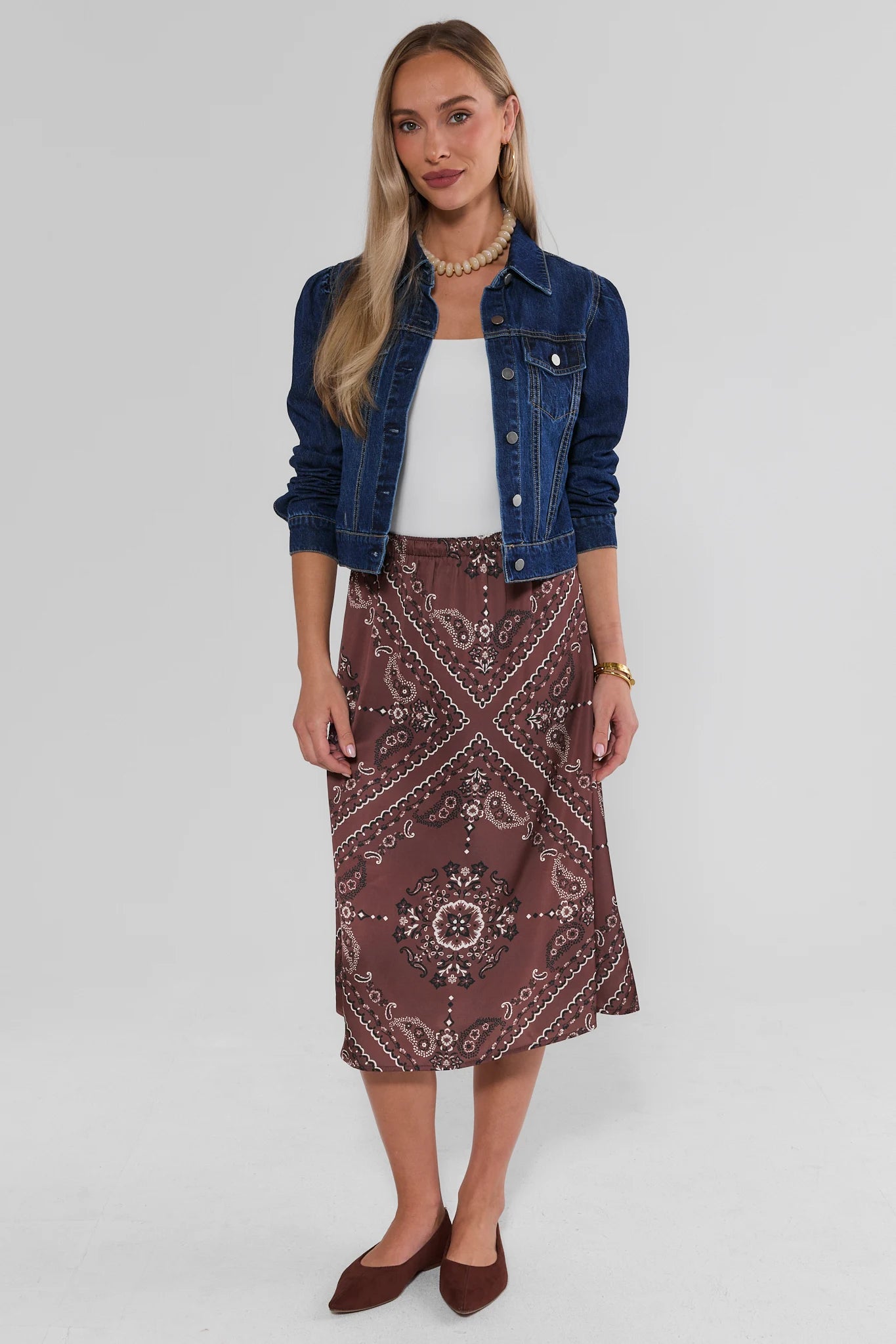 A woman modeling the Brown Bandana Print Midi Skirt with a white top, a blue denim jacket, and brown pointed flats, shot against a light gray studio background.